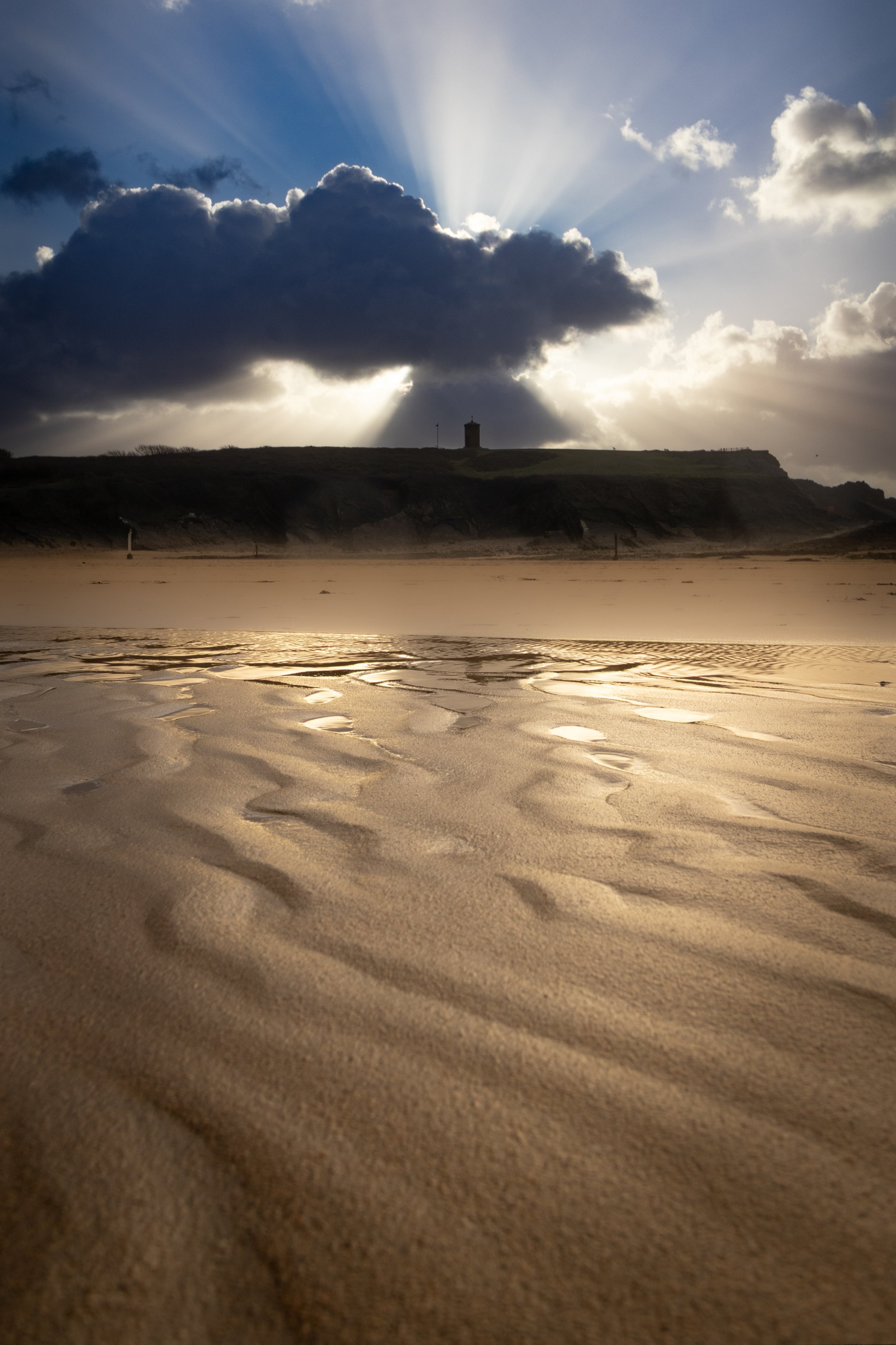 Sun rays breaking through clouds over a sandy beach with rippled sand and a grassy cliffs in the background.