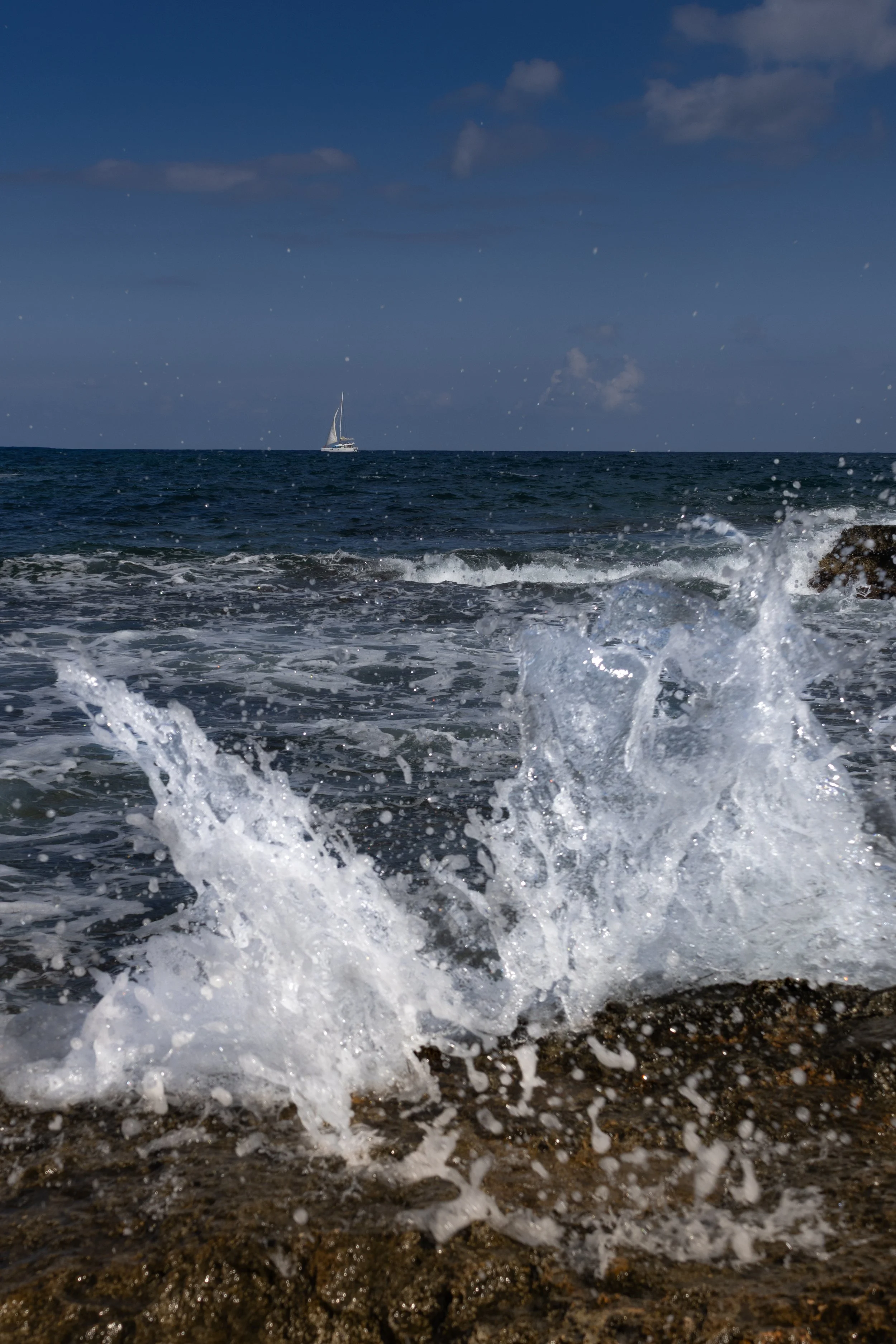 A sailboat in the distance on the ocean, with waves crashing on rocks in the foreground and a partly cloudy sky above.
