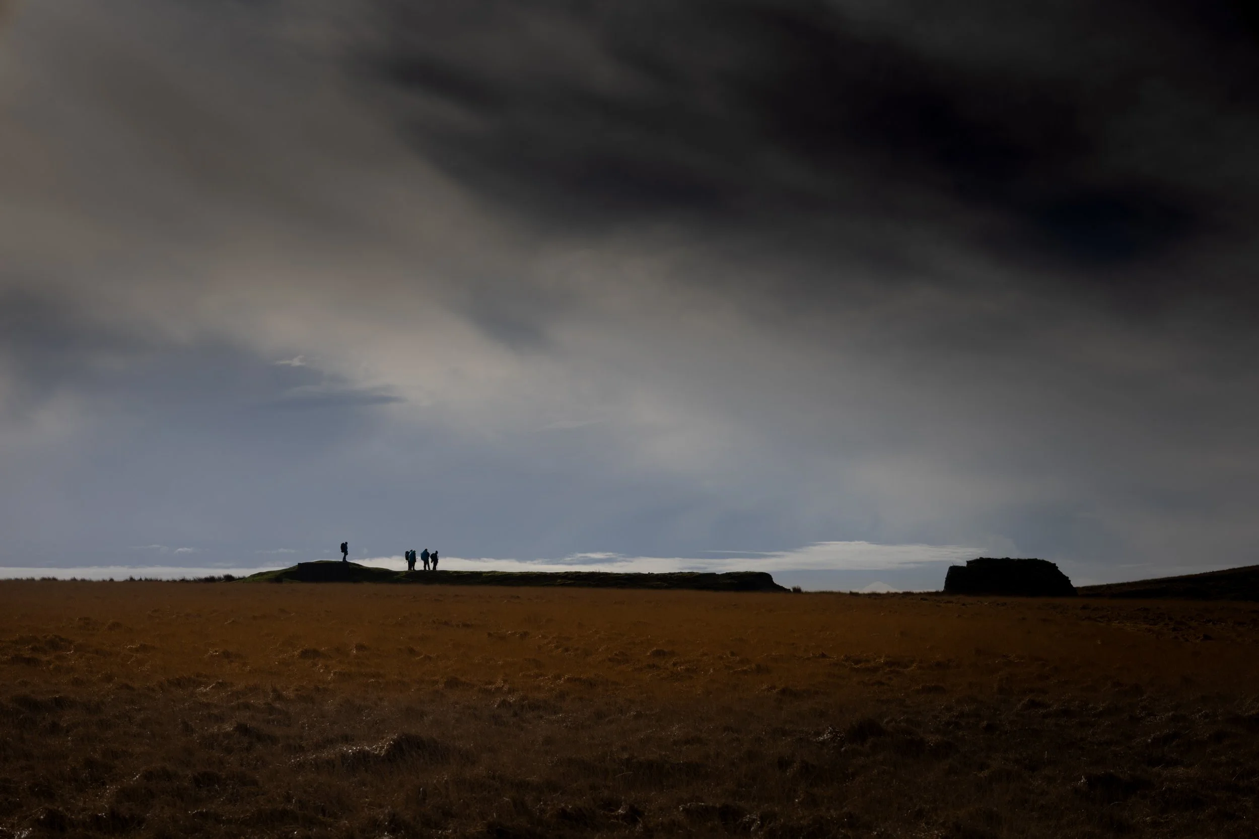 Group of people standing on a hilltop under a cloudy dark sky, with a vast open field below.
