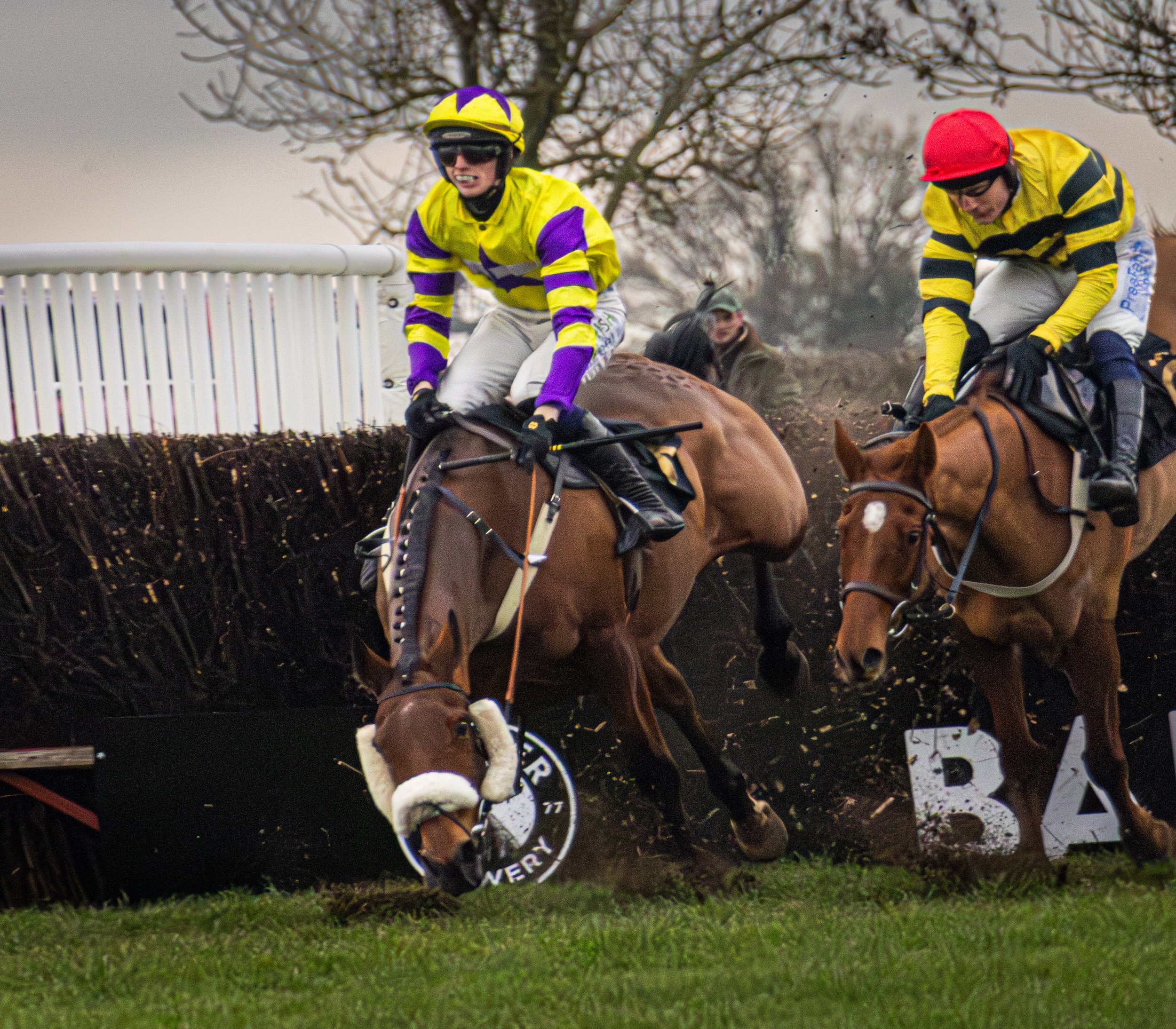 Two jockeys on horseback jumping over a hurdle in a steeplechase race, with a spectator watching in the background.