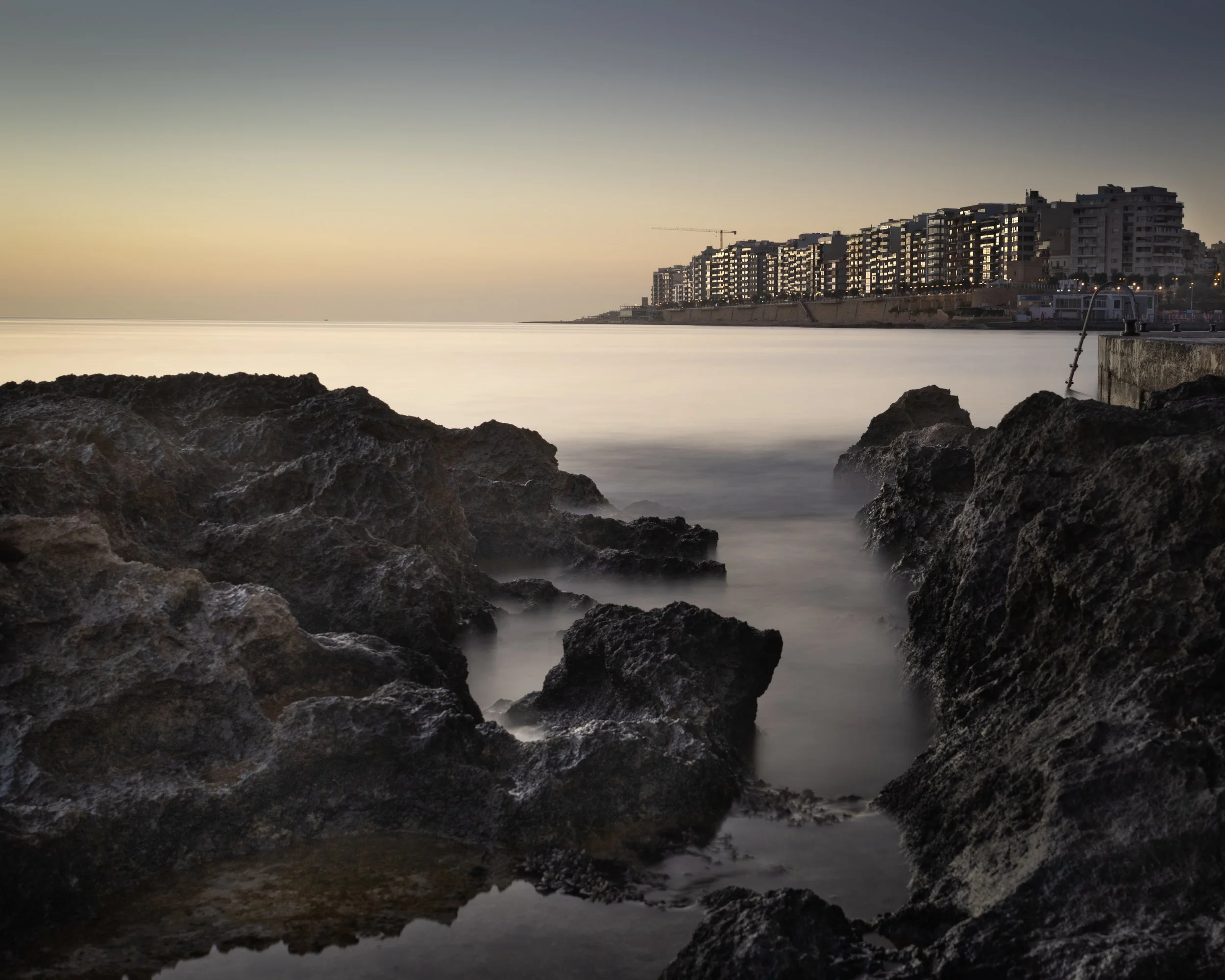 Sunset over a rocky shoreline with calm water and a cityscape in the background.