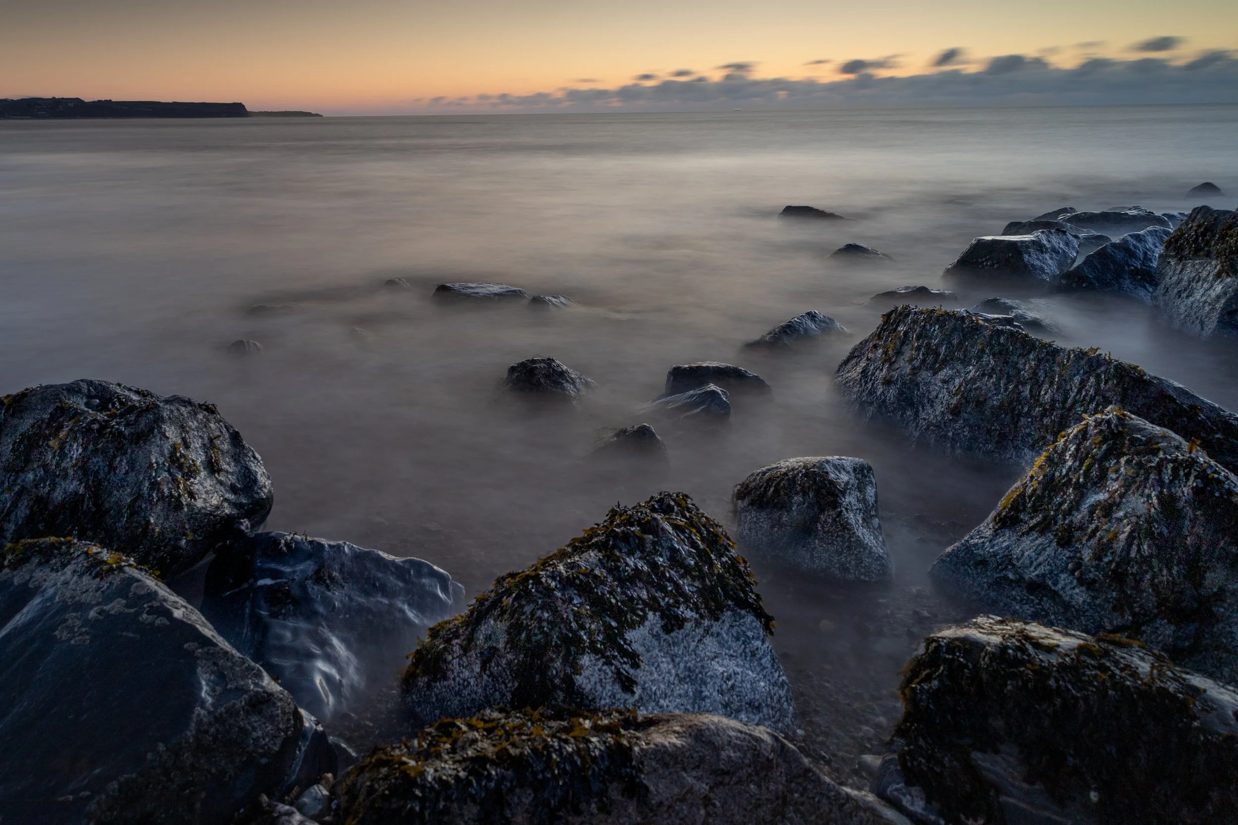 Rocky shoreline at dusk with calm ocean water and a colorful sky with clouds.