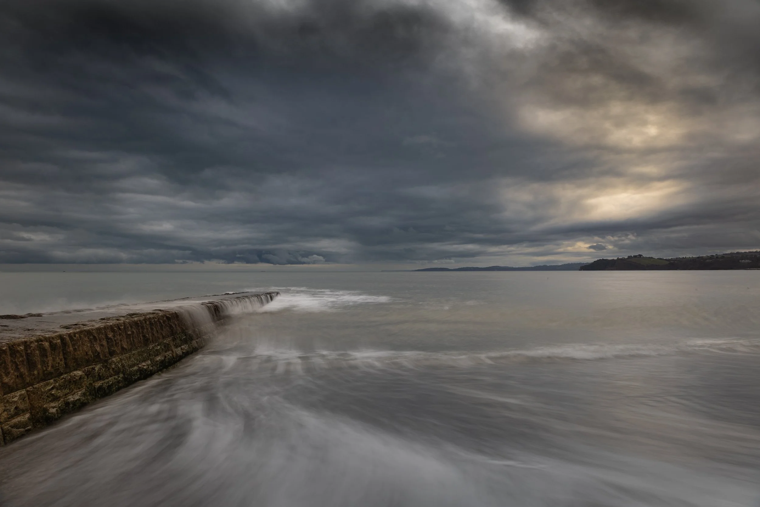 Overcast sky over a rocky sea wall with waves crashing and a distant coastline.