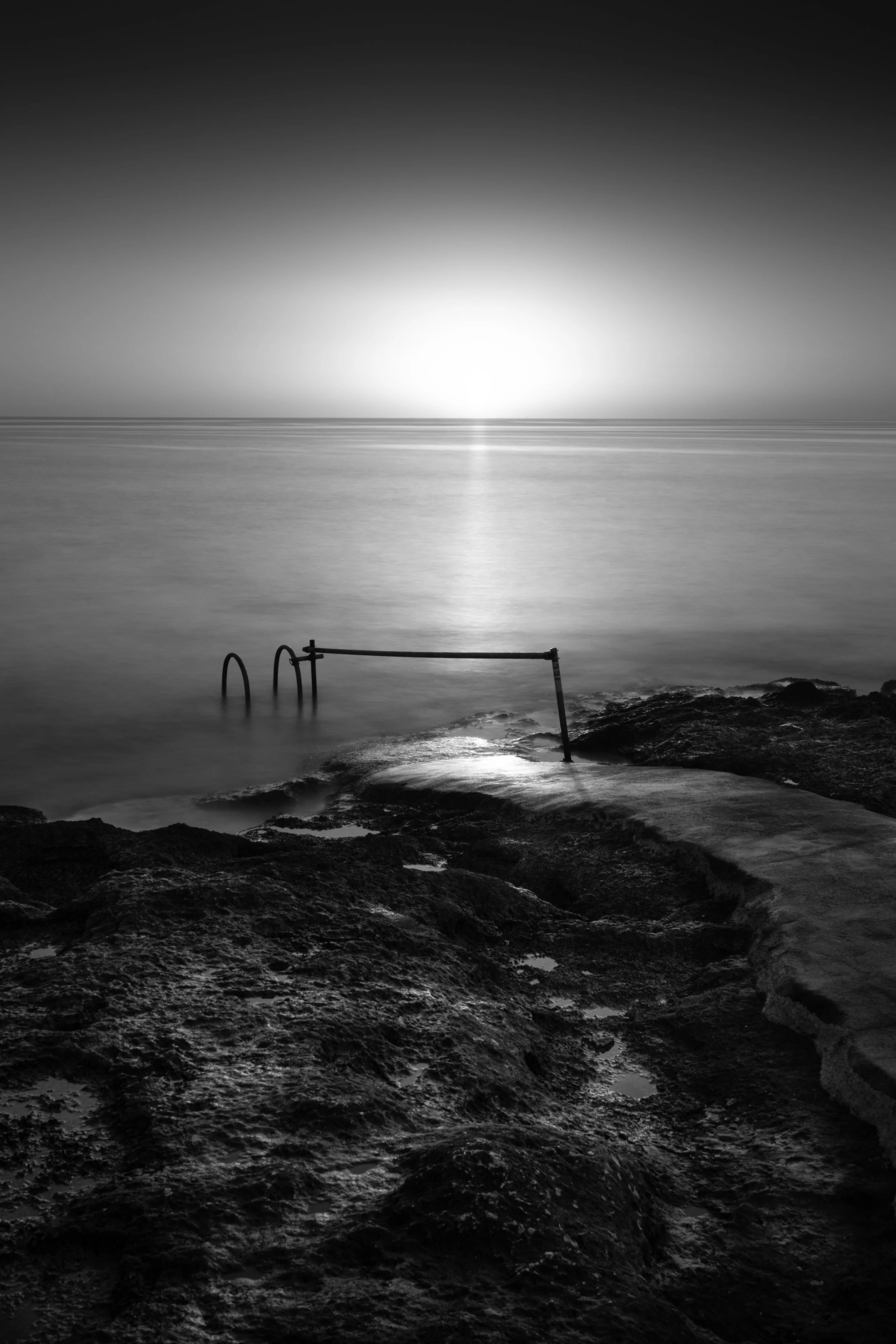 Black and white photo of a rocky seashore with a metal ladder partially submerged in water, leading into the ocean, with a setting or rising sun on the horizon.