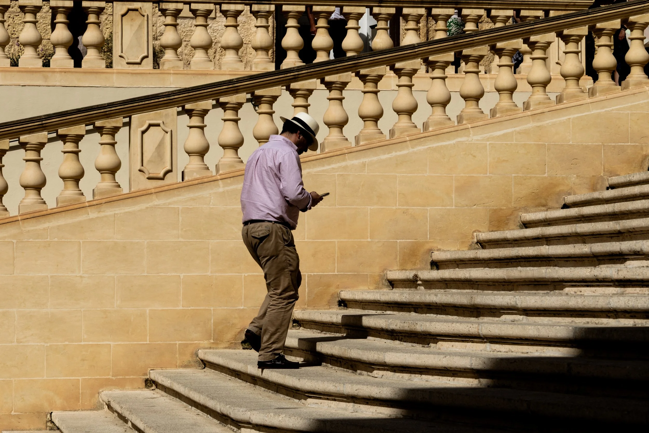 Man wearing a hat and pink shirt ascending outdoor stone stairs while looking at his phone.