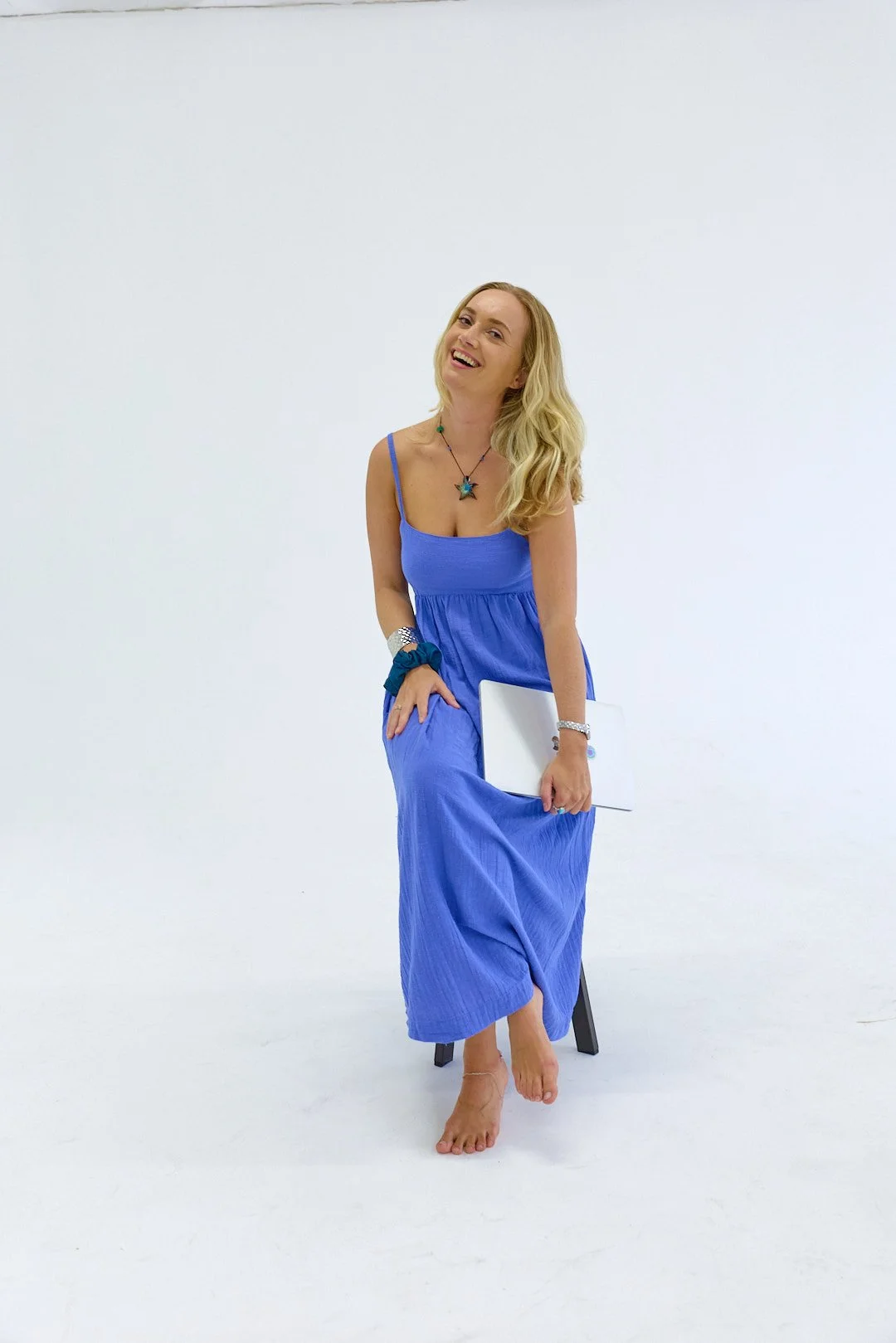 A woman with blonde hair wearing a long blue dress, sitting on a small black stool against a plain white background, holding a white folder, and smiling.