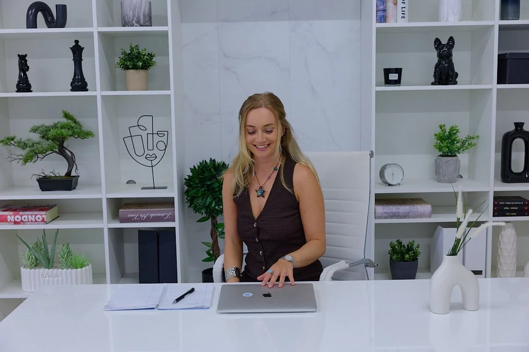 A woman sitting at a white desk in a modern office, smiling as she uses a laptop computer with notebooks and a pen in front of her, and decorative items and books on white shelves behind her.