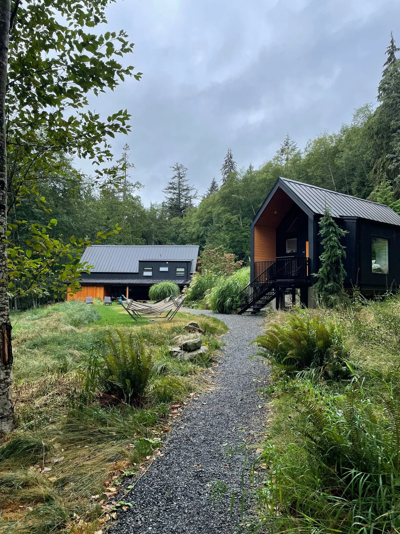 A gravel pathway leads to a modern black house with a wooden accent, set in a lush green forest with tall trees and shrubs. There is an outdoor seating area with chairs and a hammock on the grass.