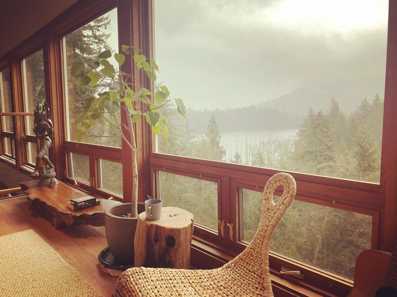 View of a forested mountainside through large windows in a cozy indoor space. Inside, there is a potted tree, a carved wooden sculpture, a coffee mug on a small tree stump, and a woven wicker chair.