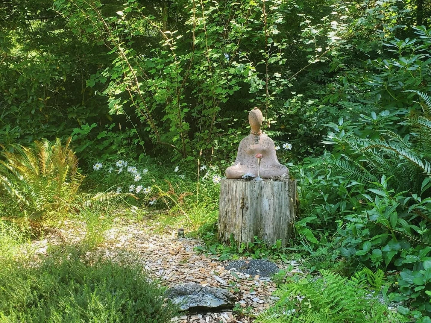 A stone sculpture of a seated person with a small mushroom in front, placed on a tree stump in a lush green garden with various plants and white flowers.
