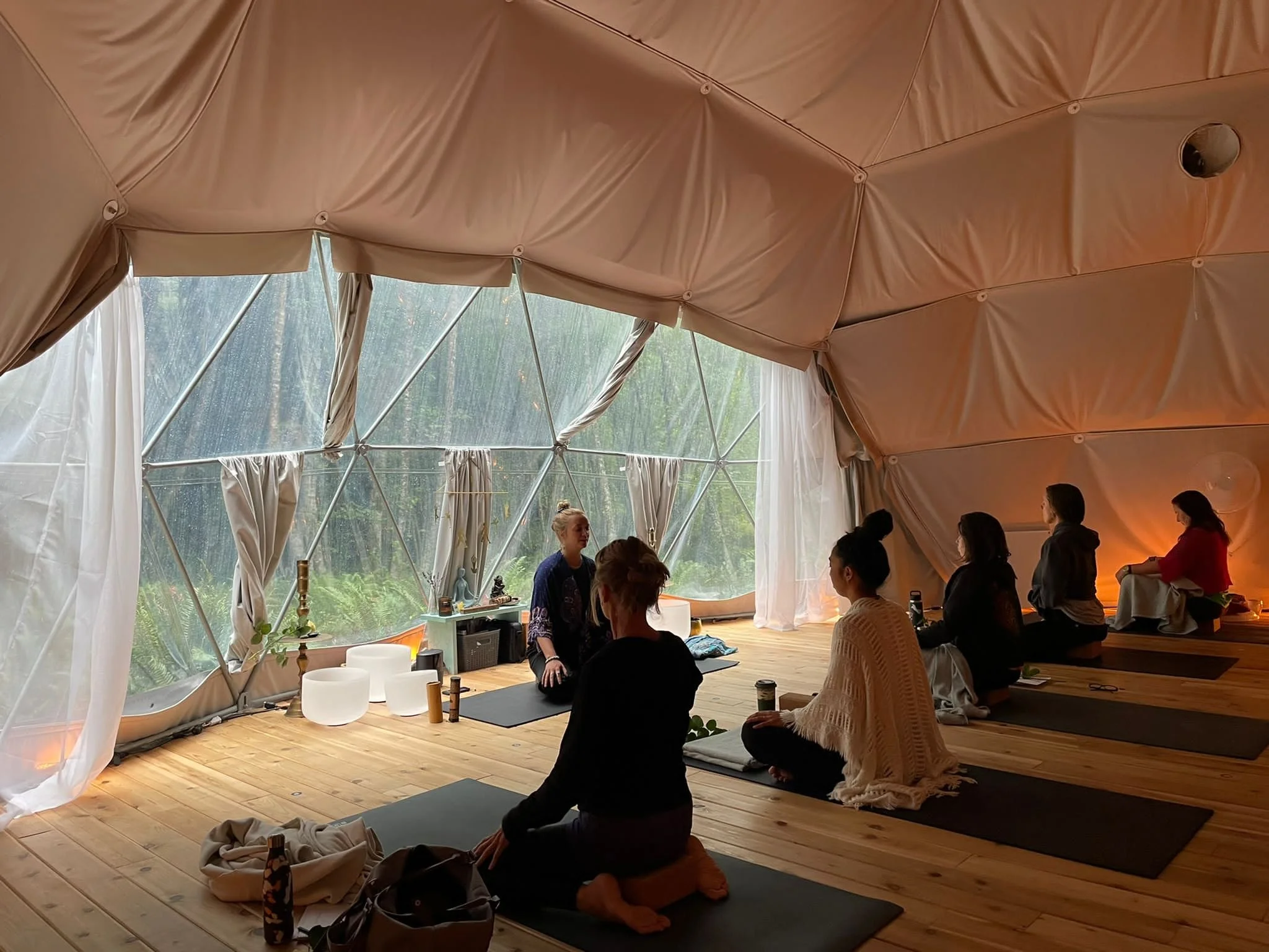 People practicing yoga or meditation inside a geodesic dome tent with large windows and curtains, wooden floor, and various mystical or decorative items.