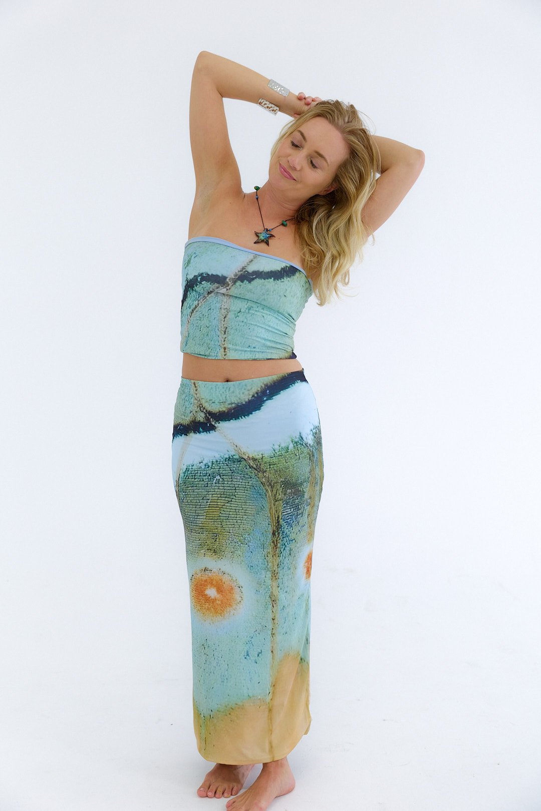 Woman wearing a matching tube top and long skirt with an abstract landscape print, arms raised behind head, standing on bare feet against a plain white background.