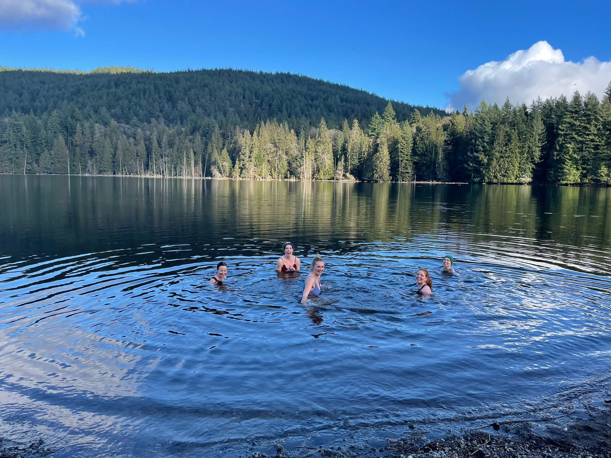 Five women swimming and enjoying a lake surrounded by trees and hills under a blue sky with clouds.