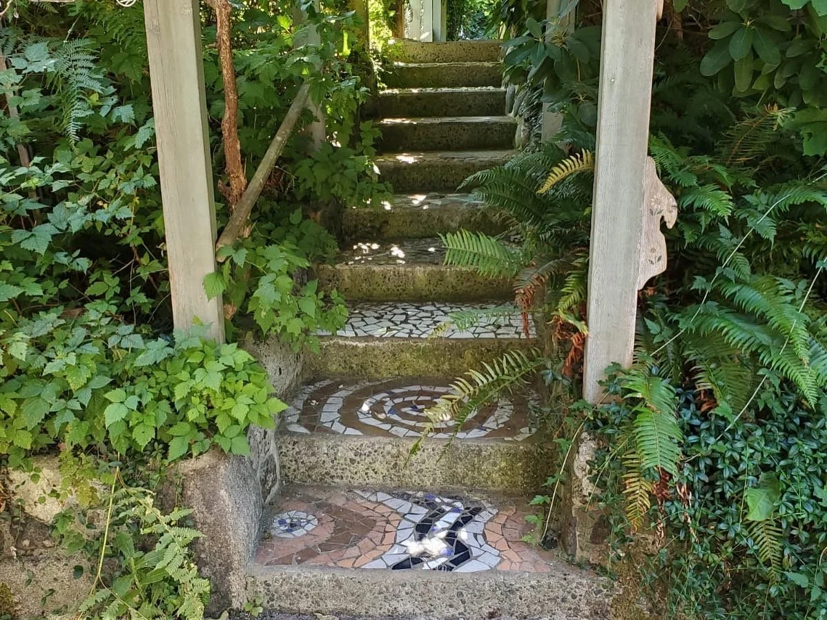 Outdoor stone staircase with mosaic tile accents, surrounded by lush green plants and foliage.