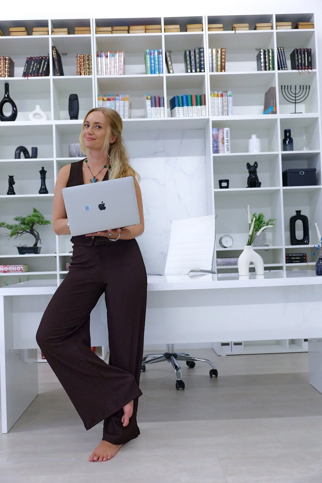 A woman stands barefoot in an office, holding a laptop, with a large white bookshelf filled with books and decorative items behind her.