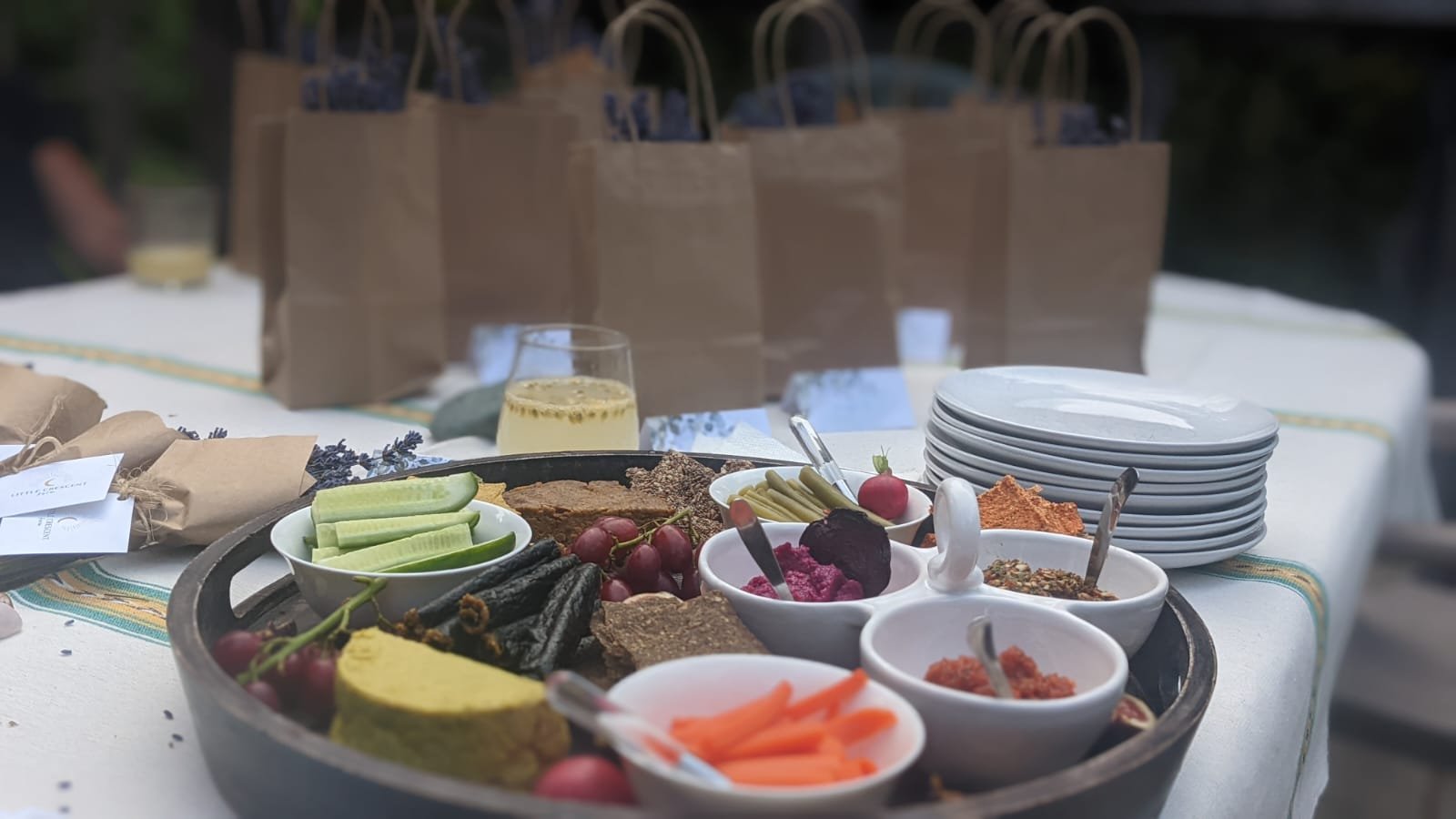 A table with assorted appetizers including sliced cucumbers, grapes, various dips and spreads in small bowls, sliced carrots, crackers, and a glass of white wine. Paper bags and stacked white plates are in the background.