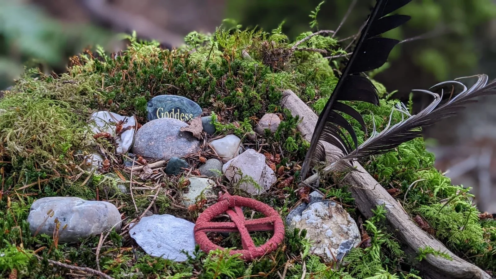 A small patch of ground with green moss, small rocks, a black feather, a red pentagram pendant, and a stone engraved with the word 'Goddess'.