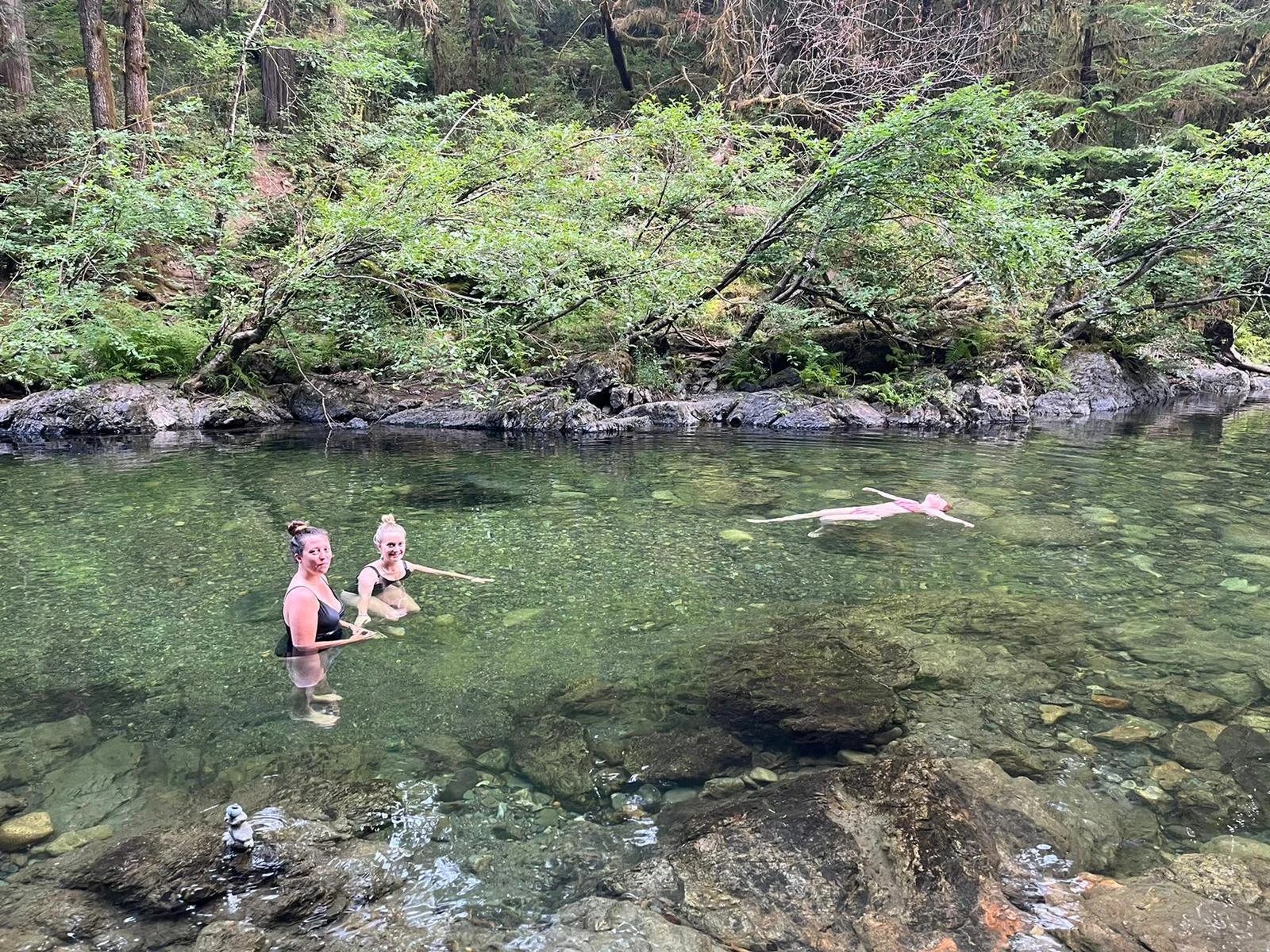 Two women in swimsuits standing in a clear river, with a dog floating on its back nearby, surrounded by lush green trees and rocks.