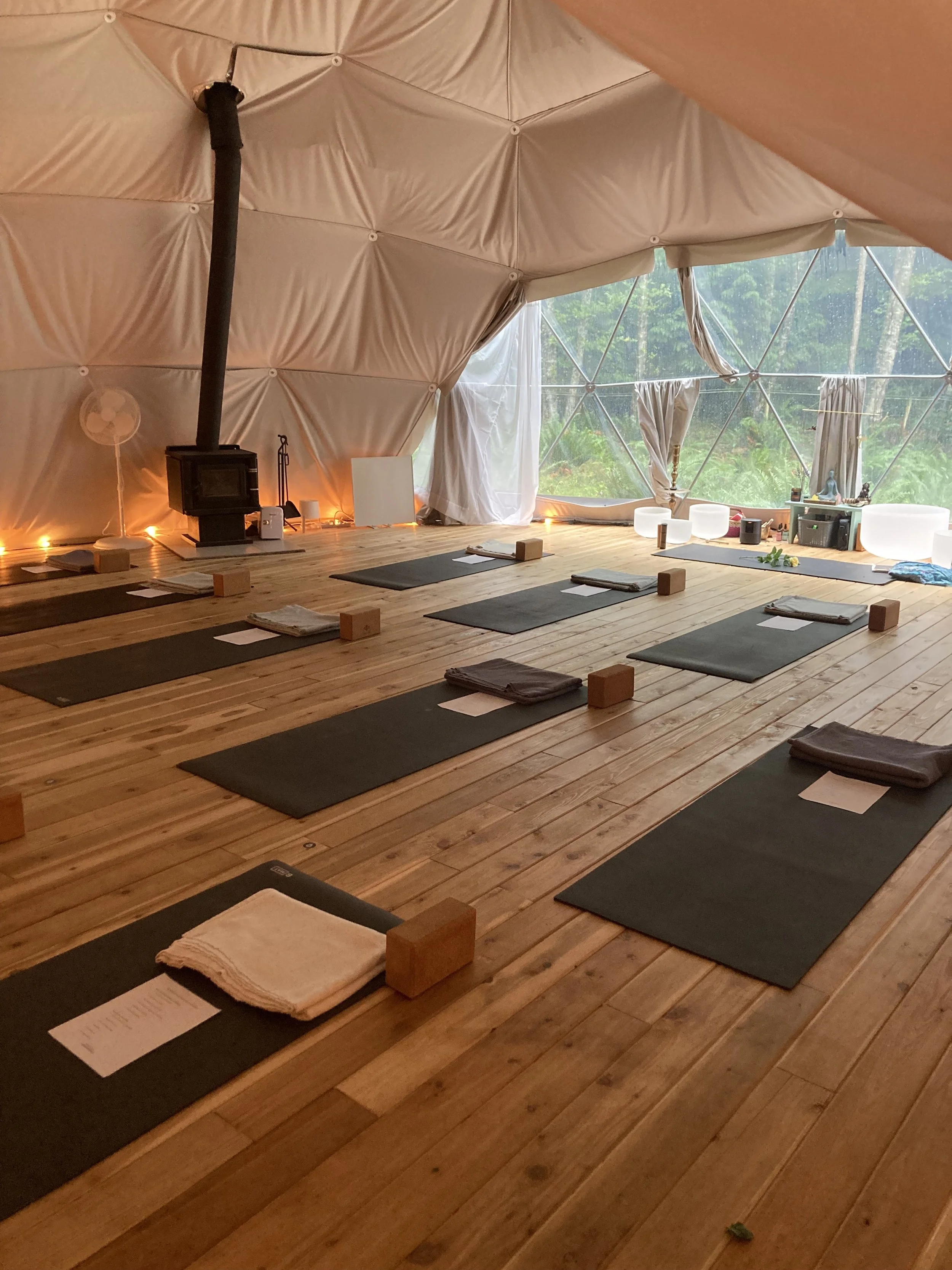 An interior of a geodesic dome with wooden floor, yoga mats, blankets, blocks, and white singing bowls, with large windows revealing lush greenery outside.