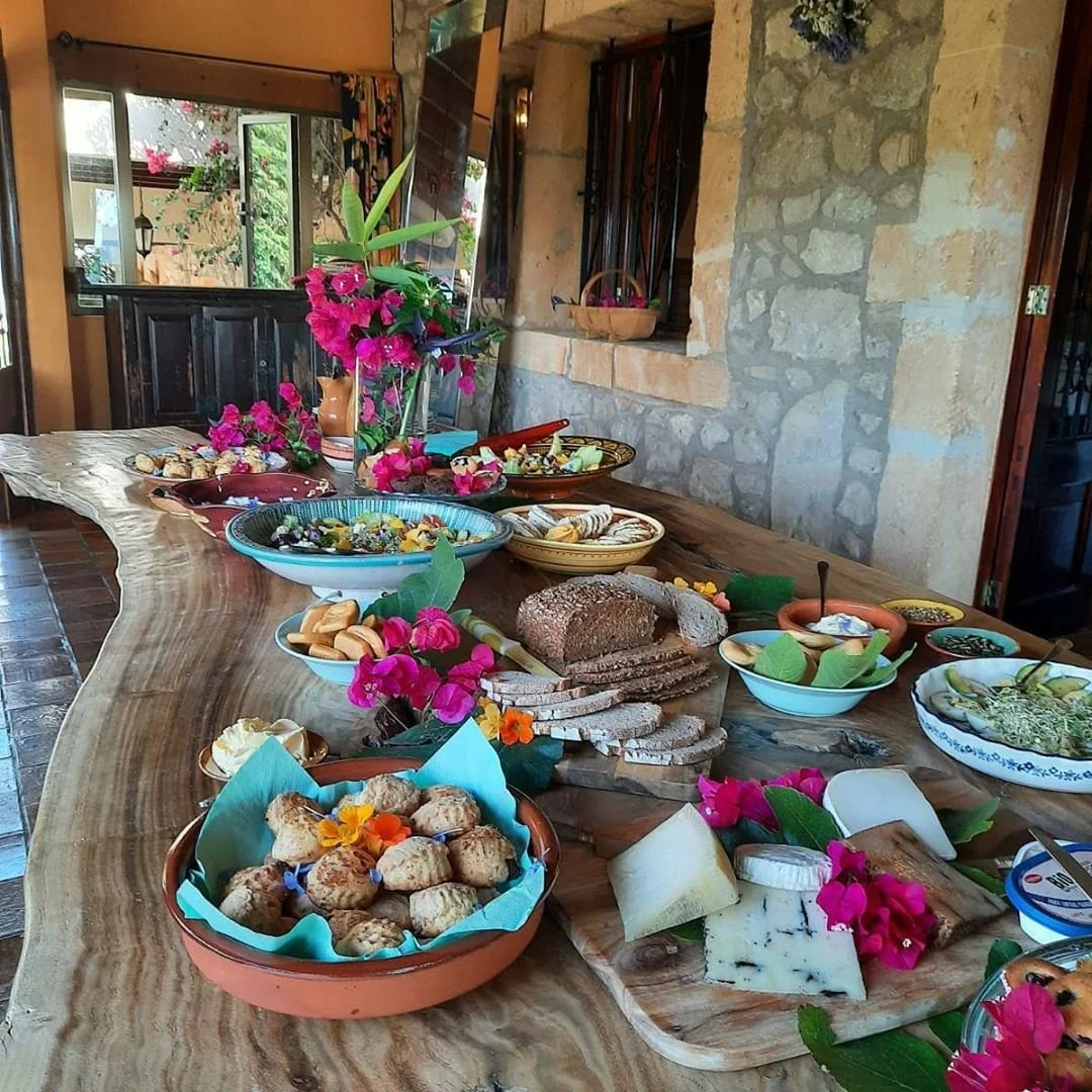 A rustic wooden table set with various dishes, cheeses, bread, and colorful flowers in a cozy room with stone and wood walls.