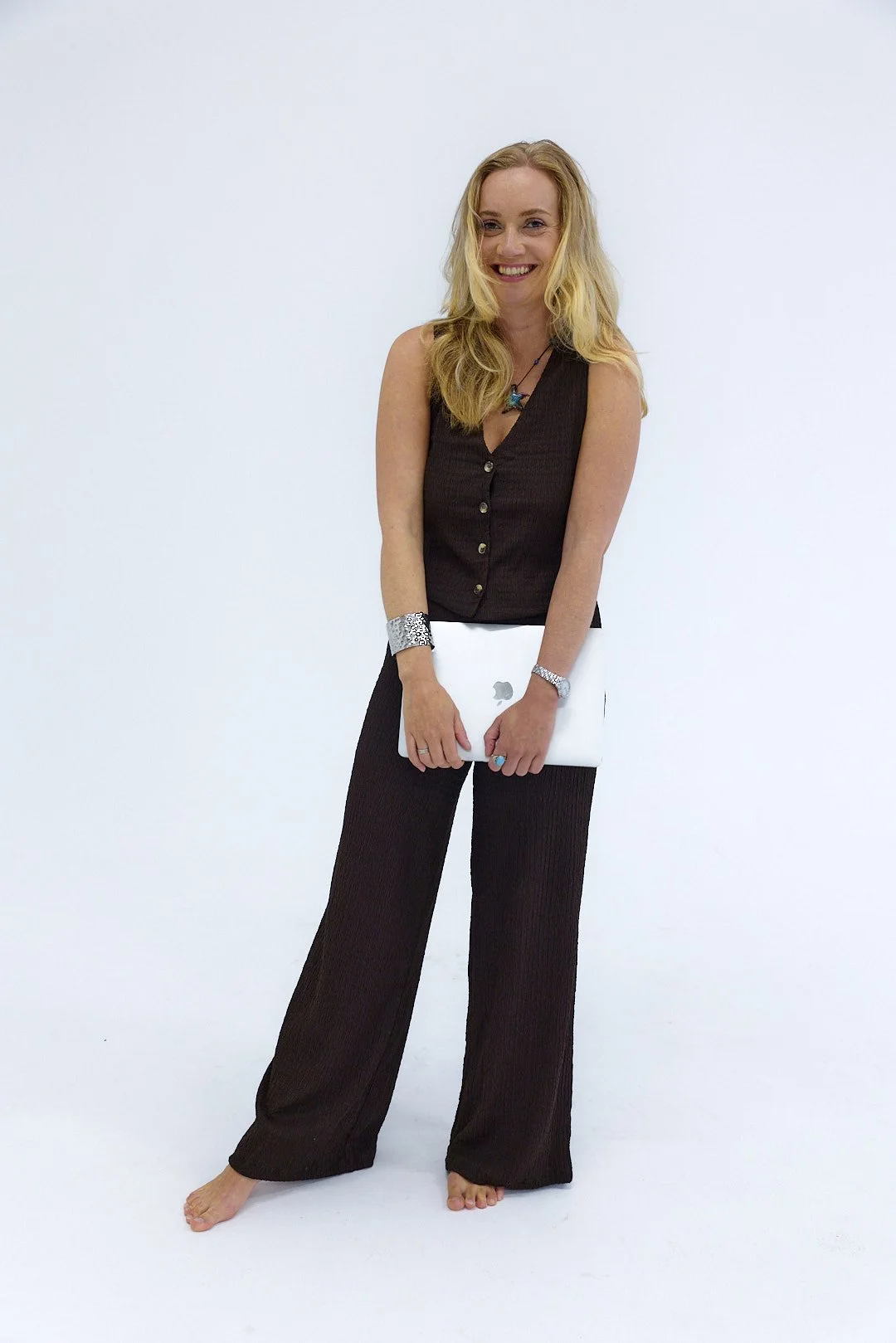 A woman with blonde hair in black sleeveless top and wide-leg pants holding a closed Apple MacBook, standing barefoot against a plain white background.