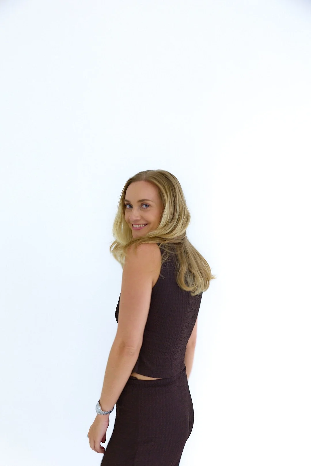 A woman with blonde hair smiling and looking over her shoulder, wearing a sleeveless dark brown dress, standing against a plain white background.