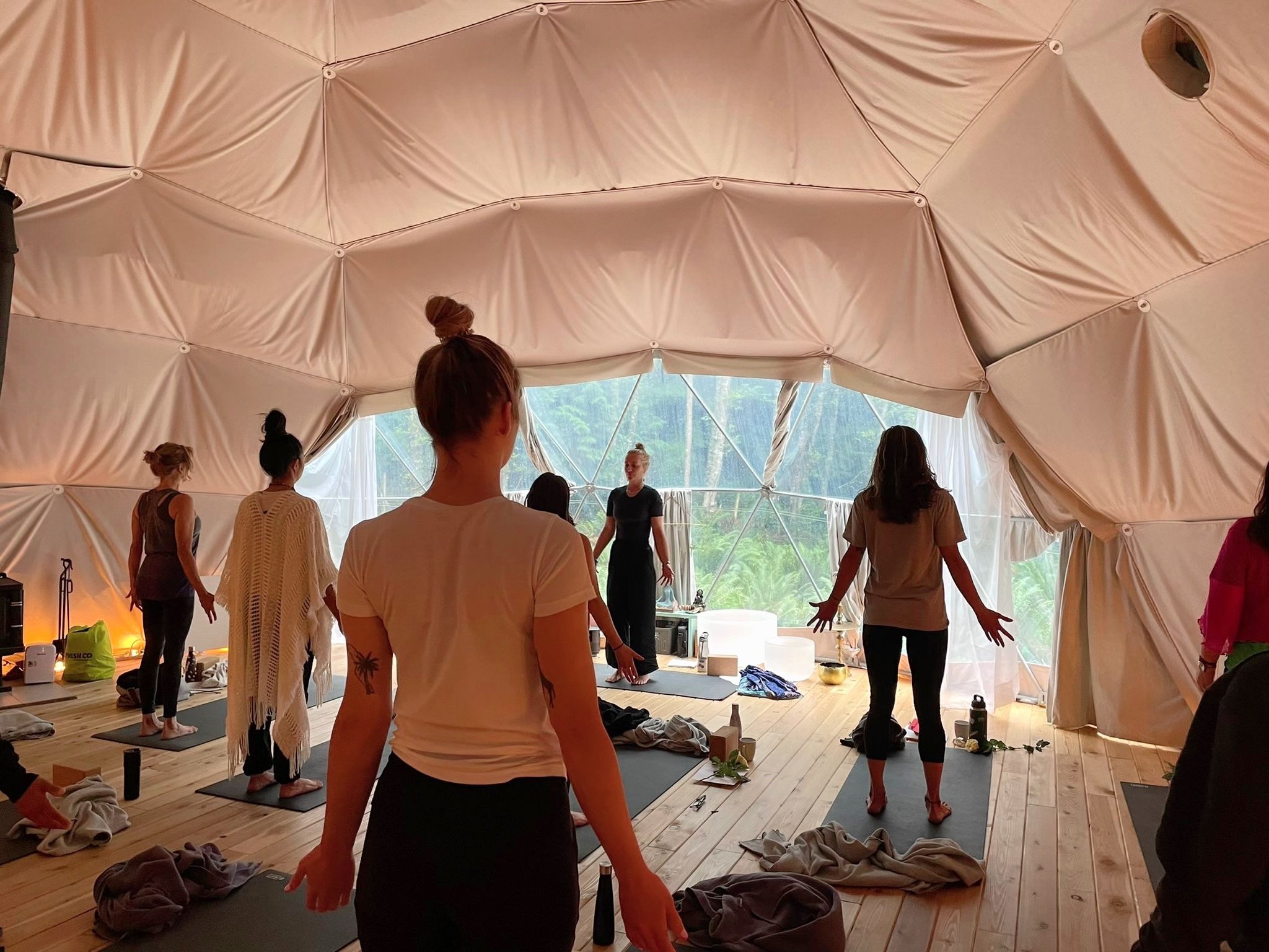 People participating in a yoga or meditation class inside a spacious tent with draped fabric on the ceiling and large windows showing outdoor greenery.