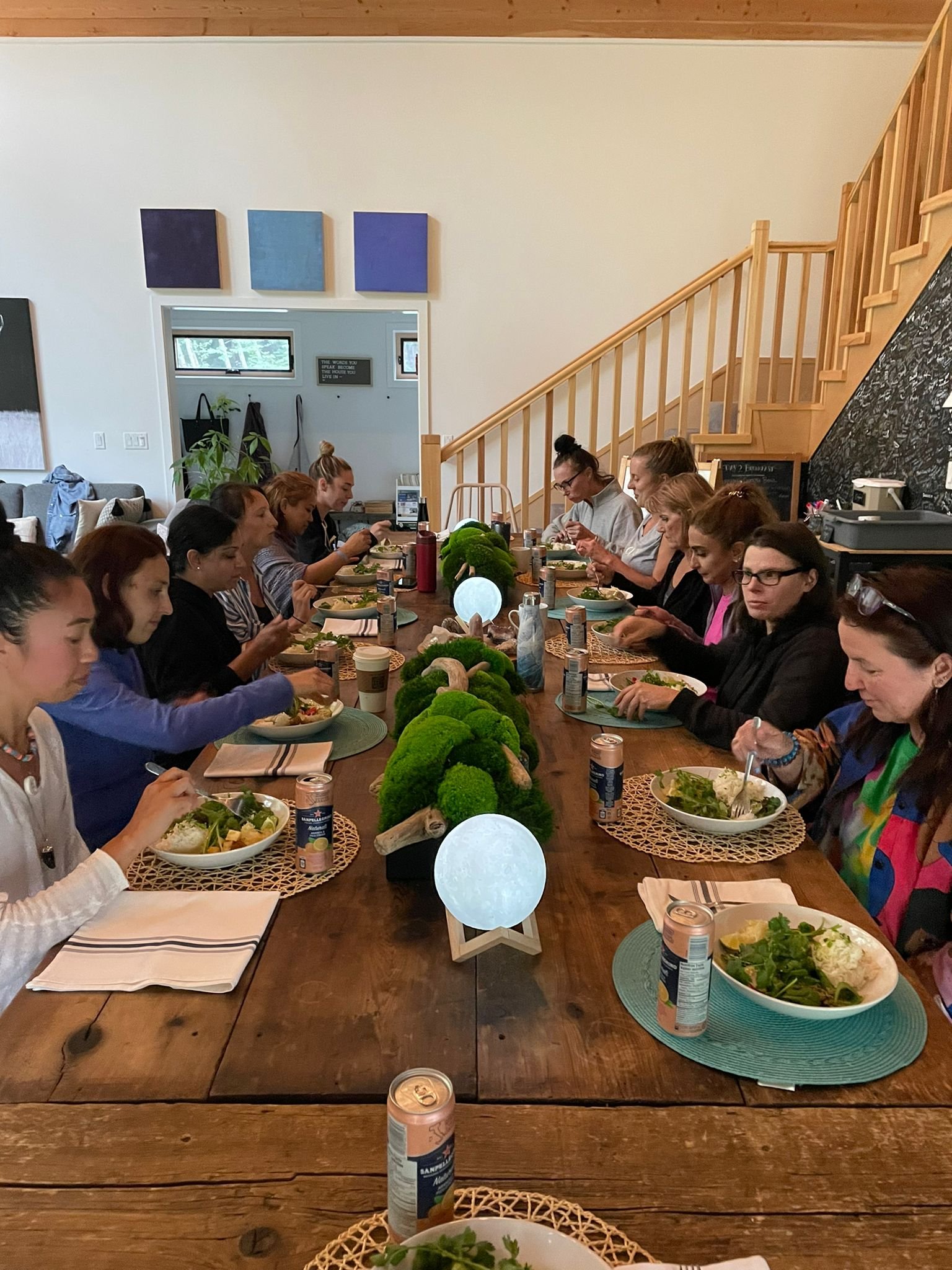 A group of women sitting around a long wooden dining table, eating salad and enjoying a meal together in a bright, modern home with a staircase and wall art in the background.