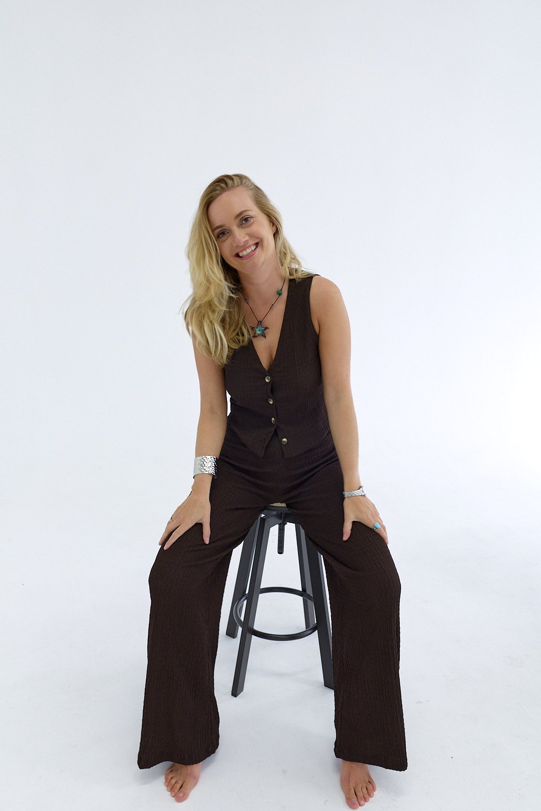 A woman with blonde hair sitting on a stool, smiling, wearing a dark sleeveless top and wide-leg pants, accessorized with necklaces, rings, and bracelets, in a white studio with a plain background.