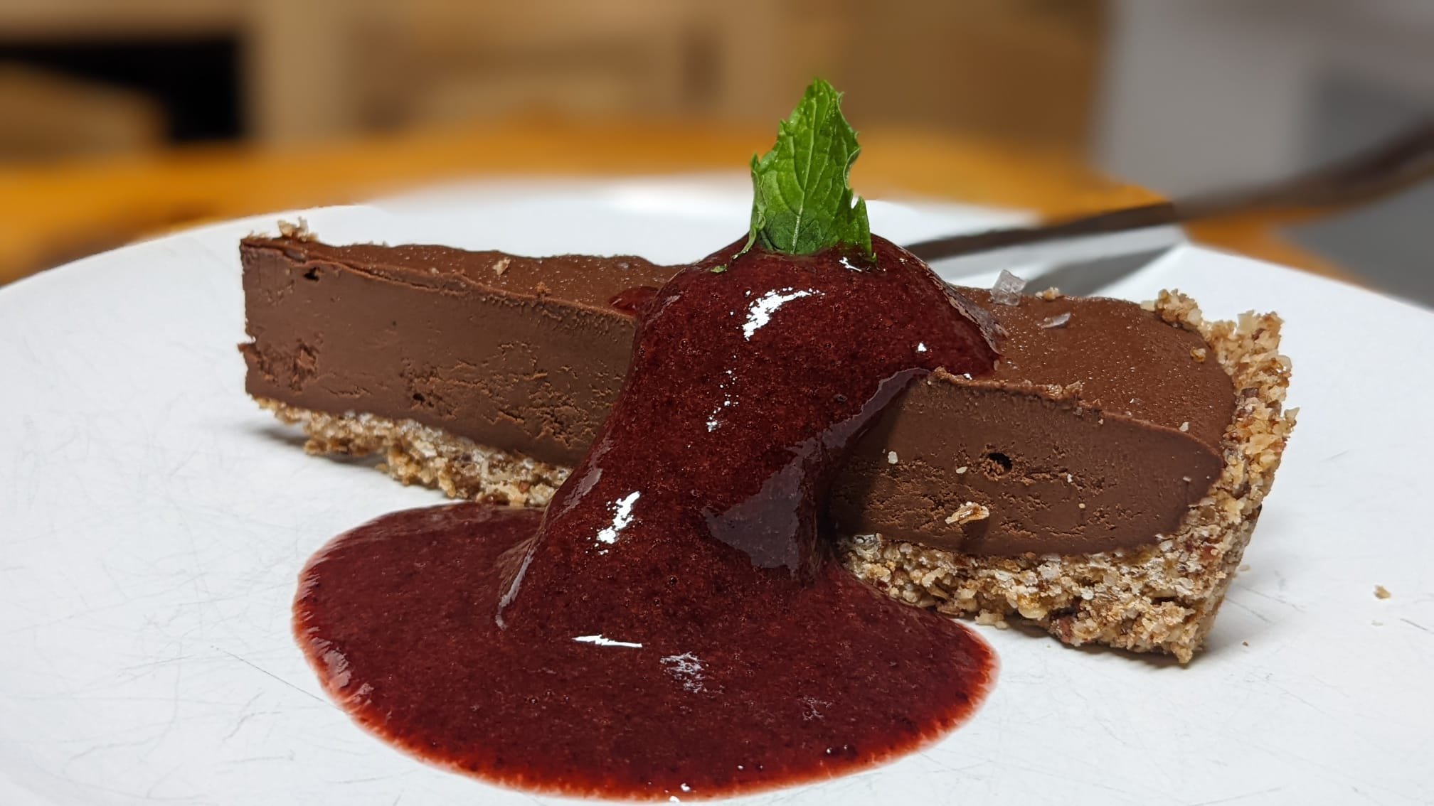 Chocolate cheesecake with a graham cracker crust, topped with a red berry sauce and garnished with a fresh mint leaf, served on a white plate.