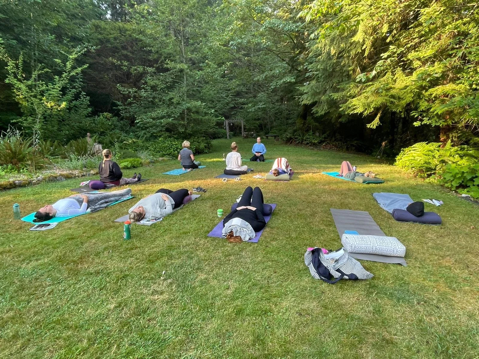 People participating in outdoor yoga or meditation session on a grassy area surrounded by trees, lying on mats with some sitting up and some lying down.