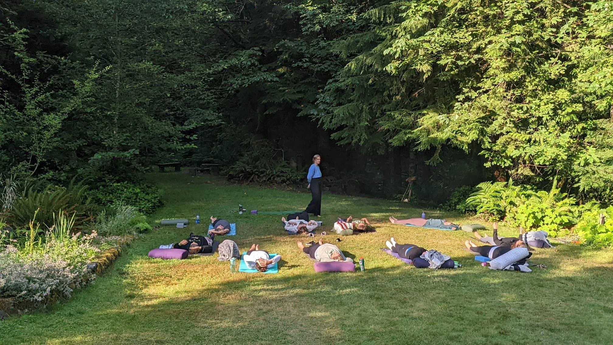 A group of people participating in an outdoor yoga or meditation class on a grassy area surrounded by lush green trees and plants, with a woman standing and leading the session.
