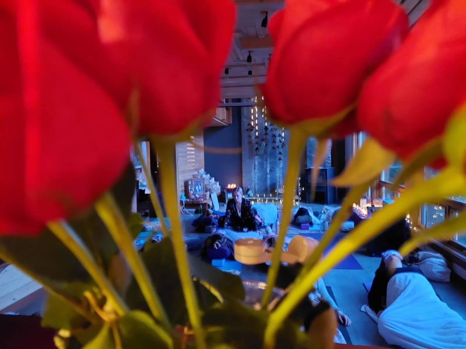 A group of people practicing yoga or meditation in a cozy, dimly lit room decorated with string lights, with a person leading the session and the participants lying on yoga mats.