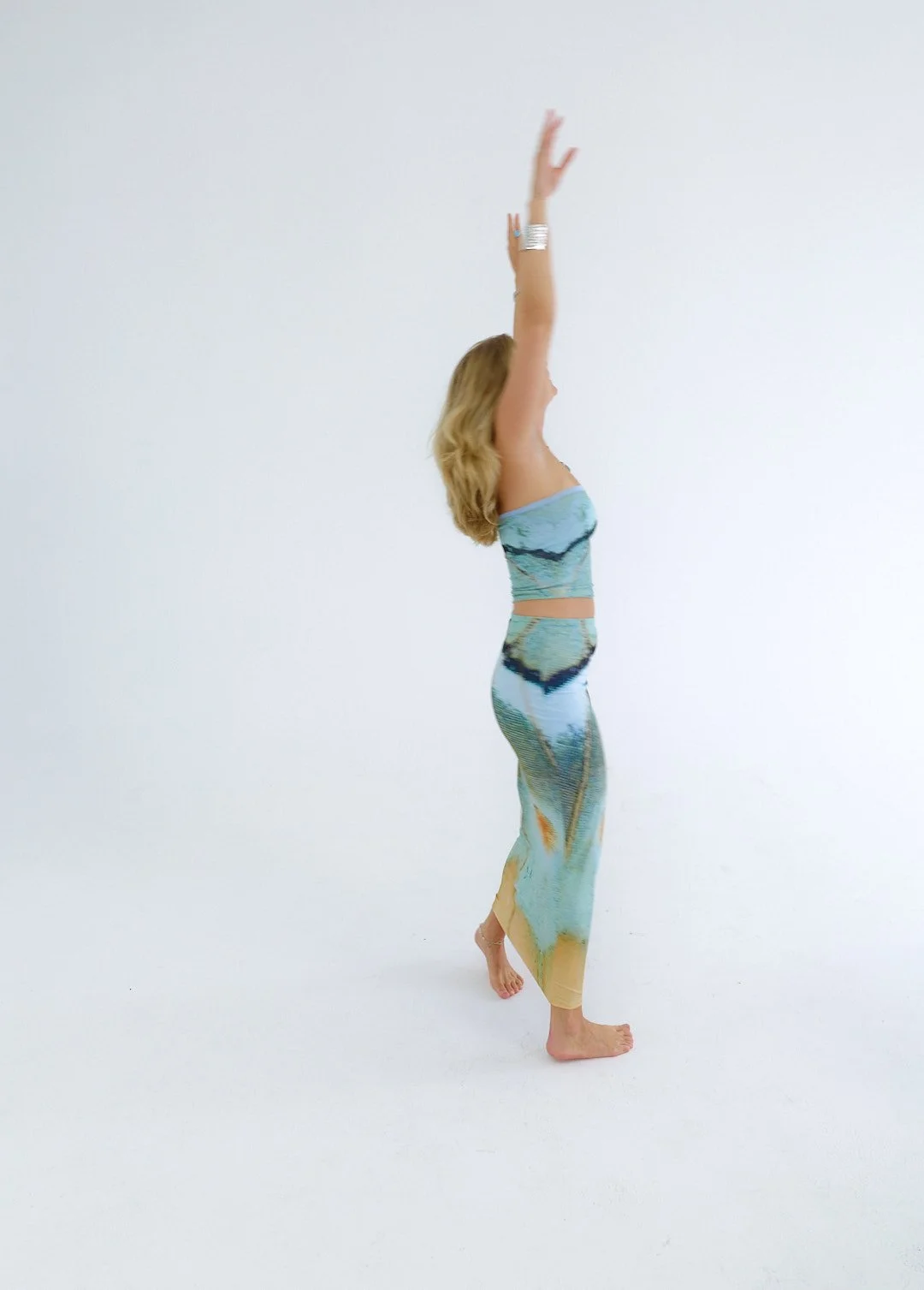 A woman with long, blonde hair standing barefoot against a plain white background, wearing a matching outfit with a landscape print, stretching with arms raised above her head.