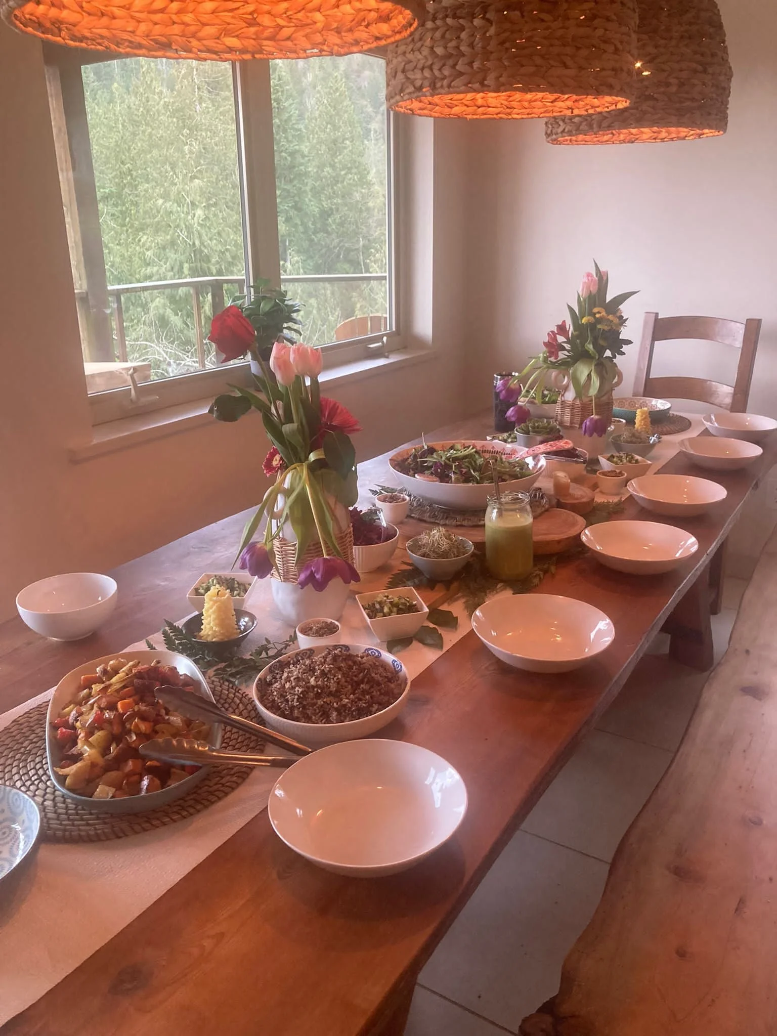 A dining table set for a meal with various bowls of food, two flower vases with flowers, and leafy decorations. Hanging lamps provide lighting, and a window shows green trees outside.