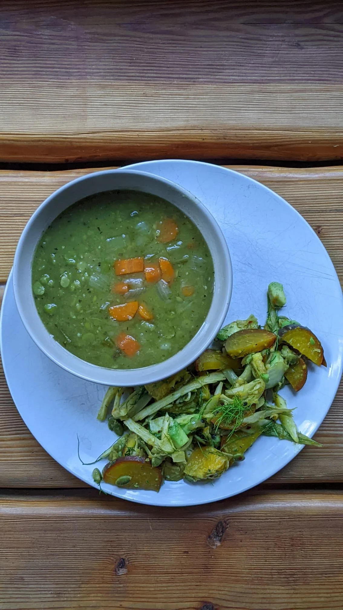 A white plate holding a bowl of green vegetable soup with carrots and a side of mixed vegetables, placed on a wooden table.