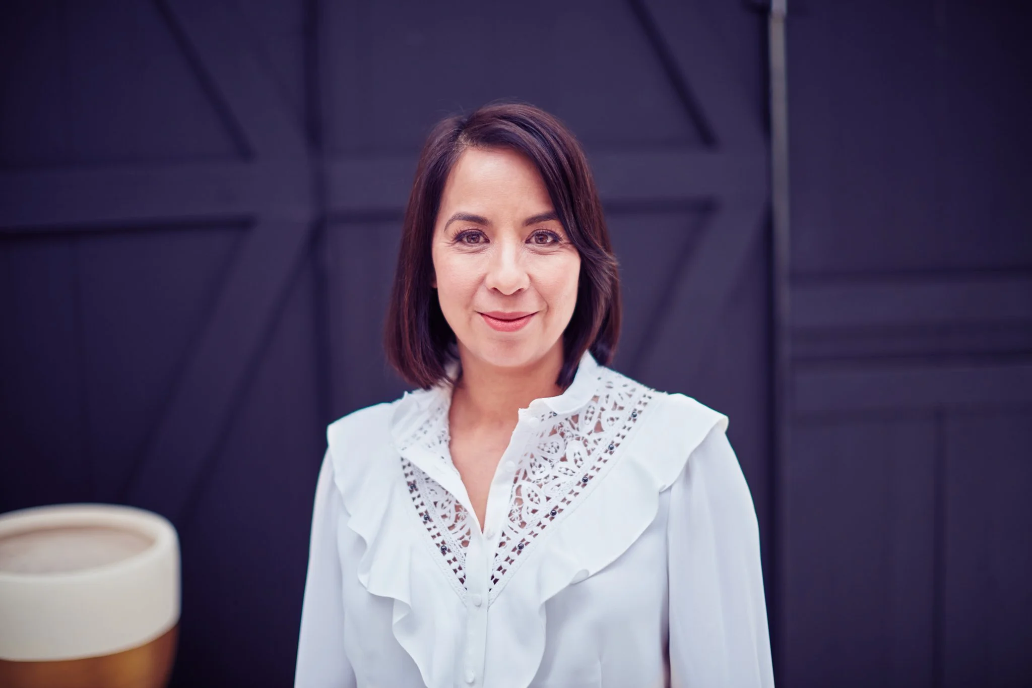 A woman with shoulder-length brown hair wearing a white blouse with lace details, standing against a black patterned wall.
