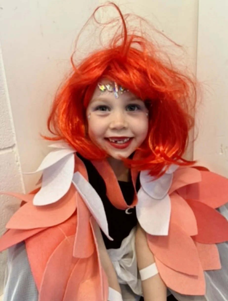 A young child wearing a bright red wig with a wild, spiky style and a costume with large pink and white petal-like layers, smiling cheerfully with face paint and small decorative stones on the forehead.