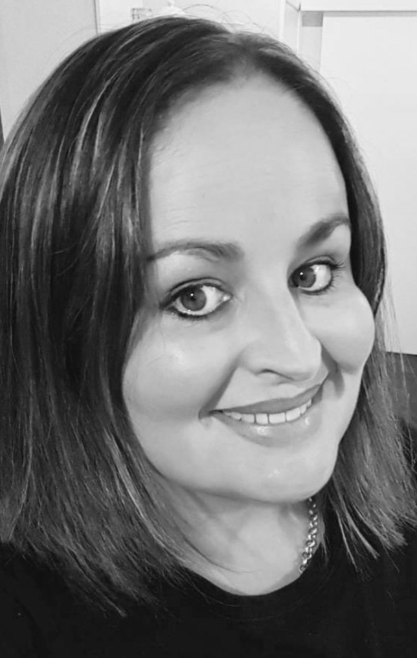 A black and white close-up portrait of a woman with shoulder-length dark hair, light eyes, and a smile, wearing a chain necklace.