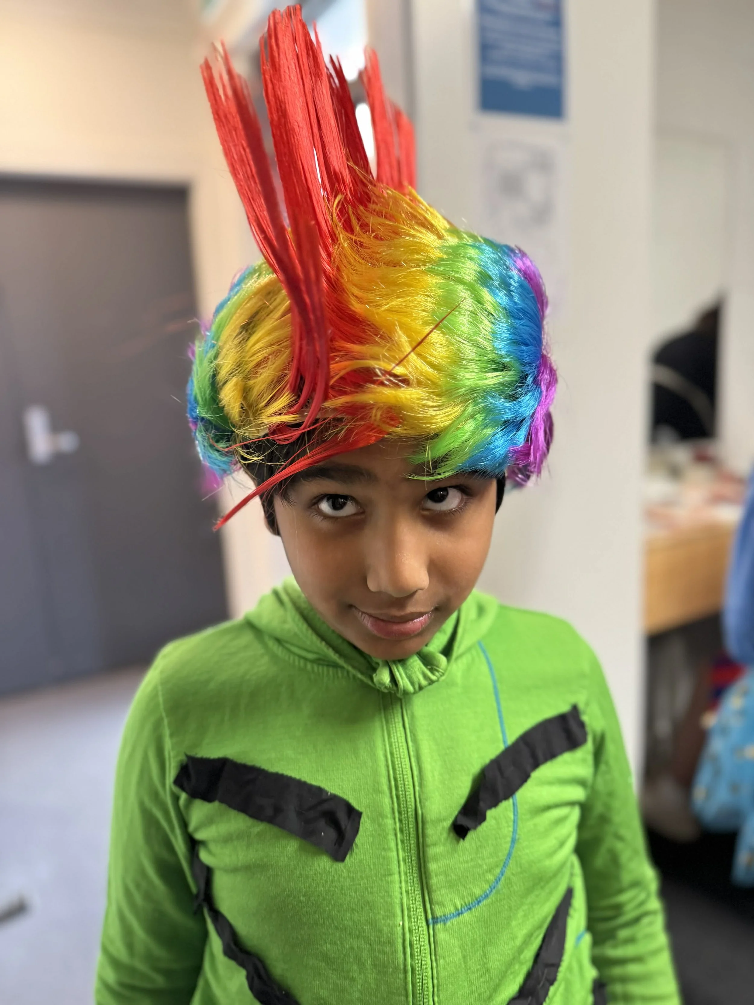 A boy wearing a bright green jacket with black accents and a zip-up front, sporting a colorful rainbow clown wig with tall red spikes, looking directly at the camera in an indoor setting.
