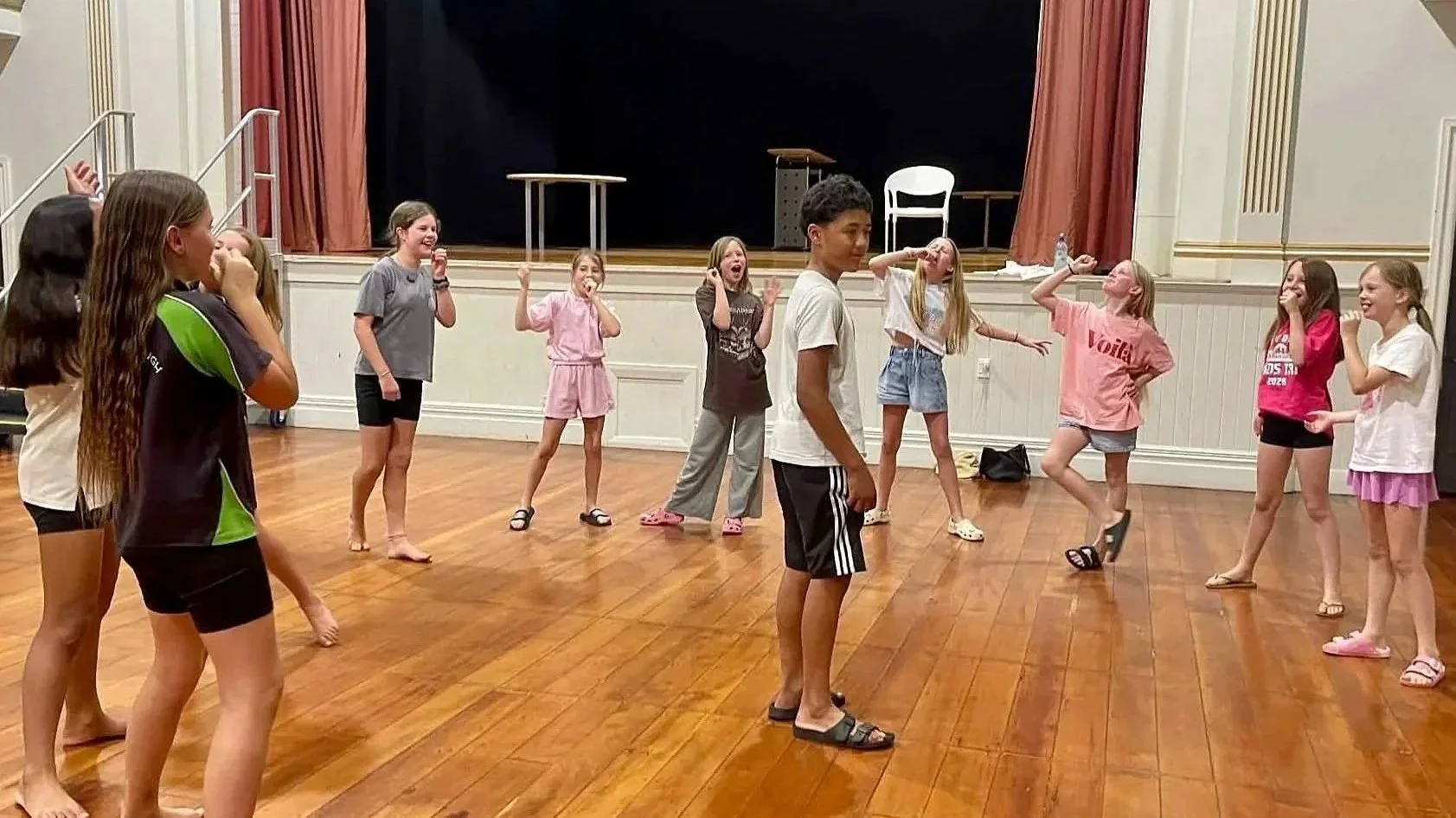 Children standing in a circle on a wooden floor in a theater room, laughing and playing.