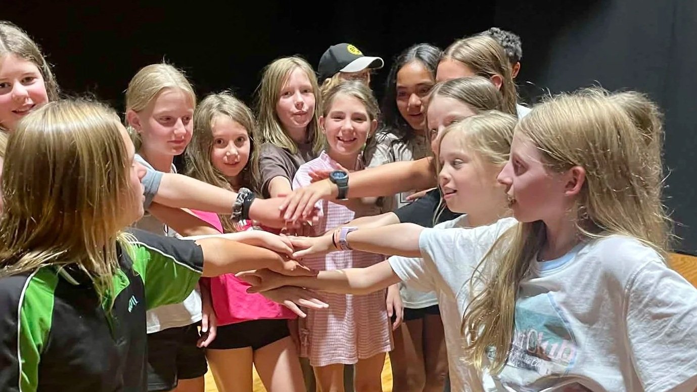 Group of young girls with their hands stacked together in the center, smiling and cheering on a stage or rehearsal space.