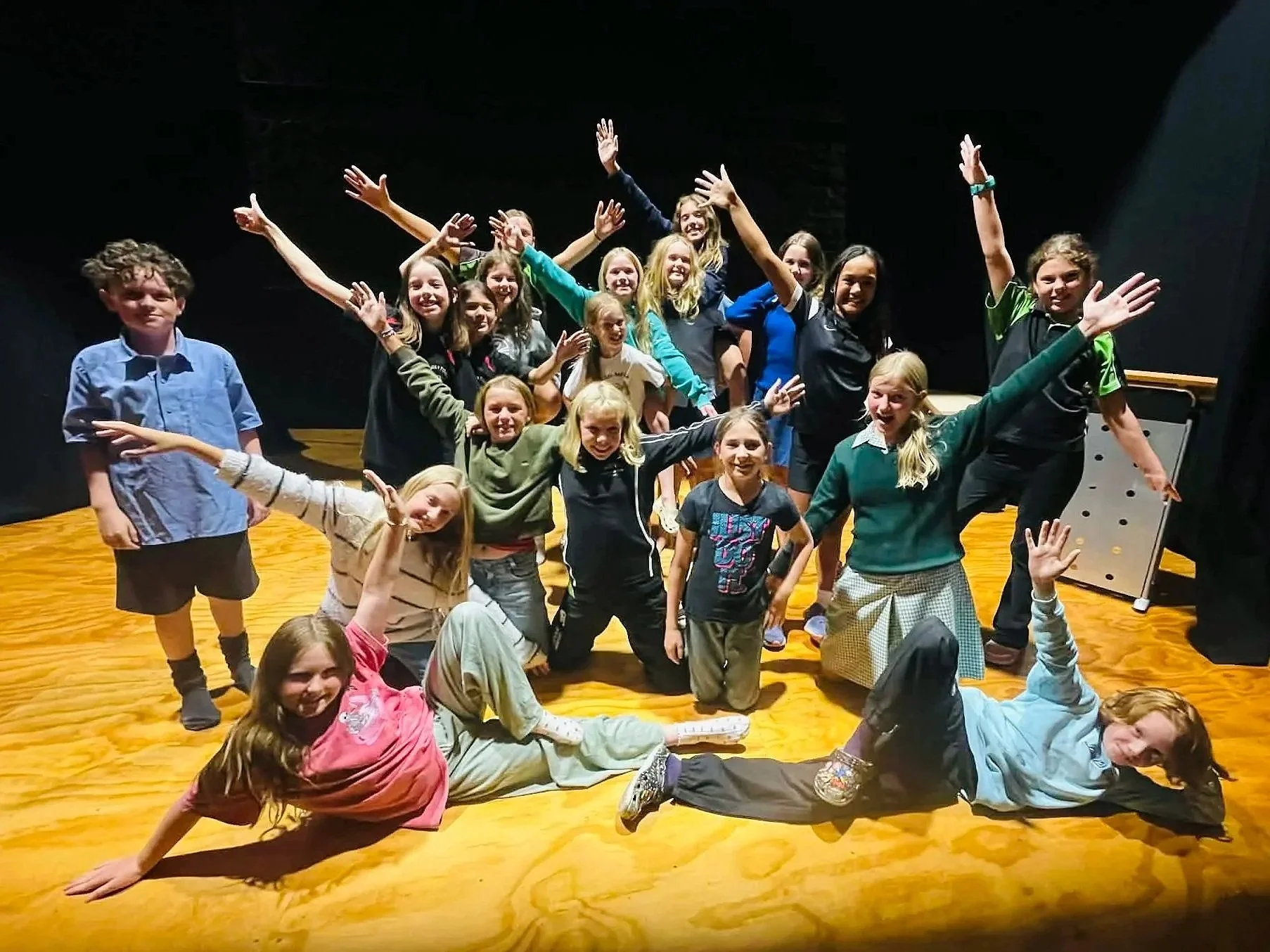 A group of children on a wooden stage, smiling and posing with their arms raised and extended in various directions.