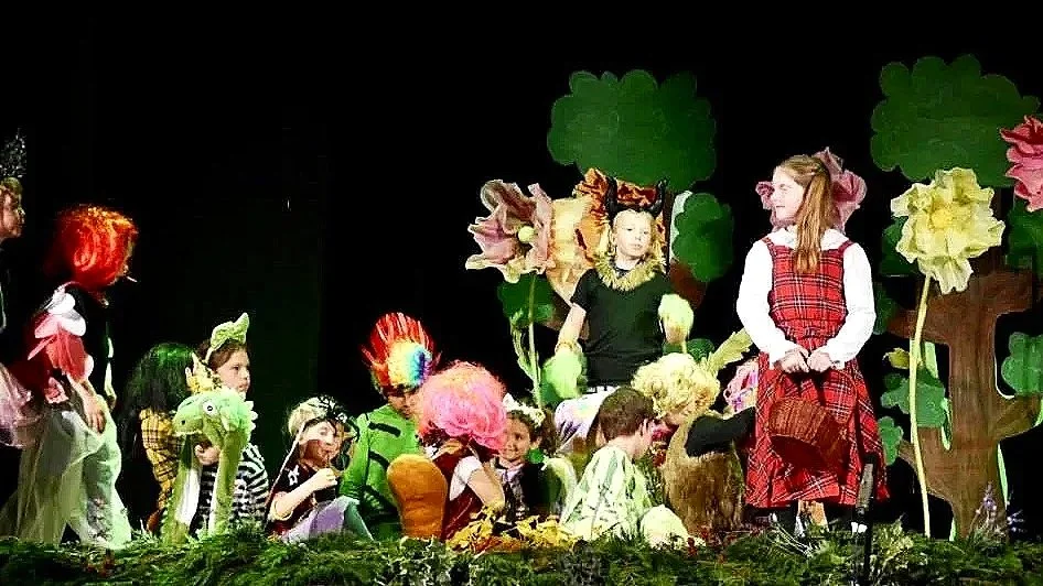 Children dressed as characters from Alice in Wonderland performing on stage with colorful oversized flowers and a forest backdrop.