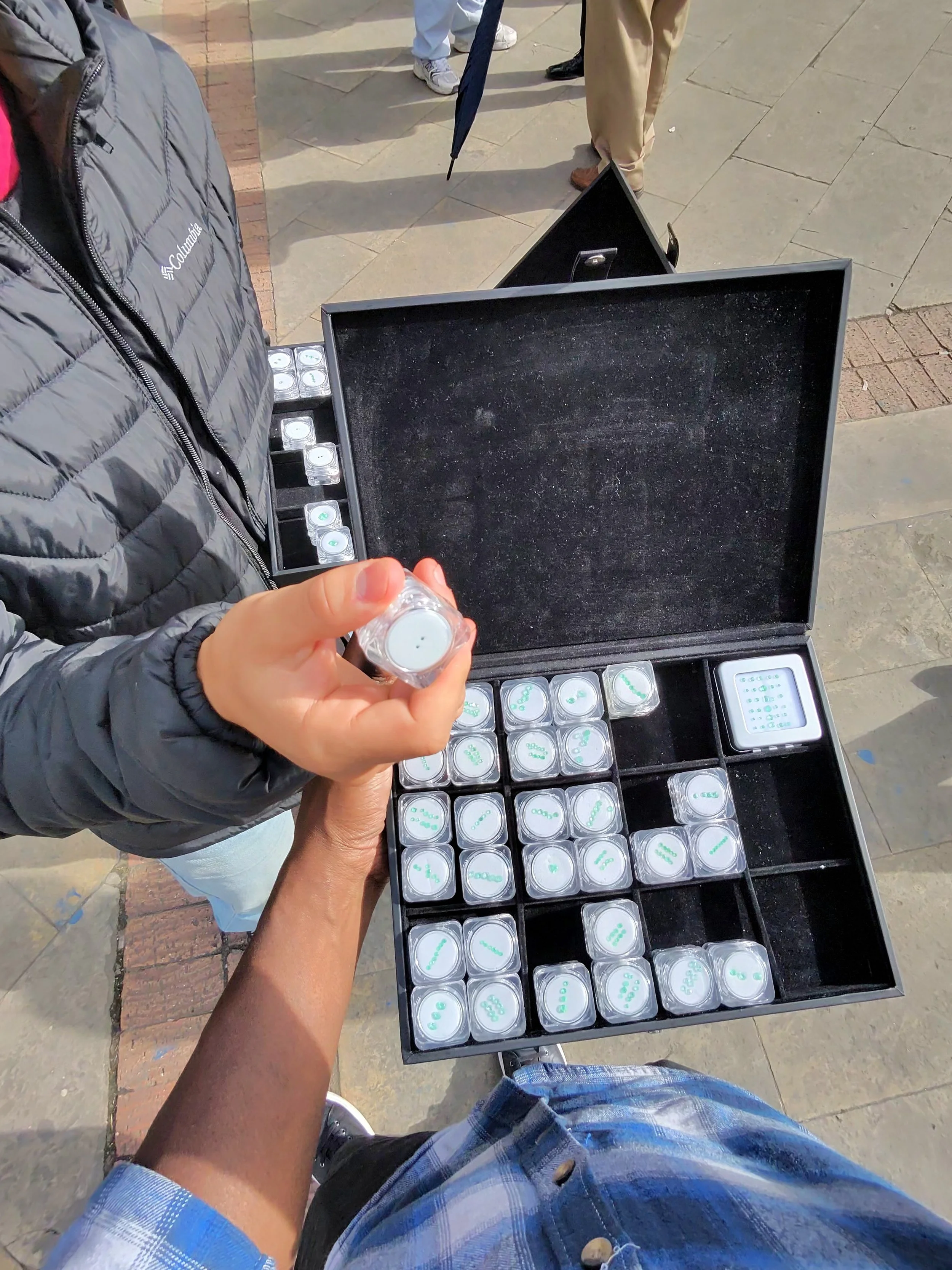 A person in shorts and a plaid shirt stands outdoors near a table filled with small containers of pills or tablets, some of which are being handed to another person.