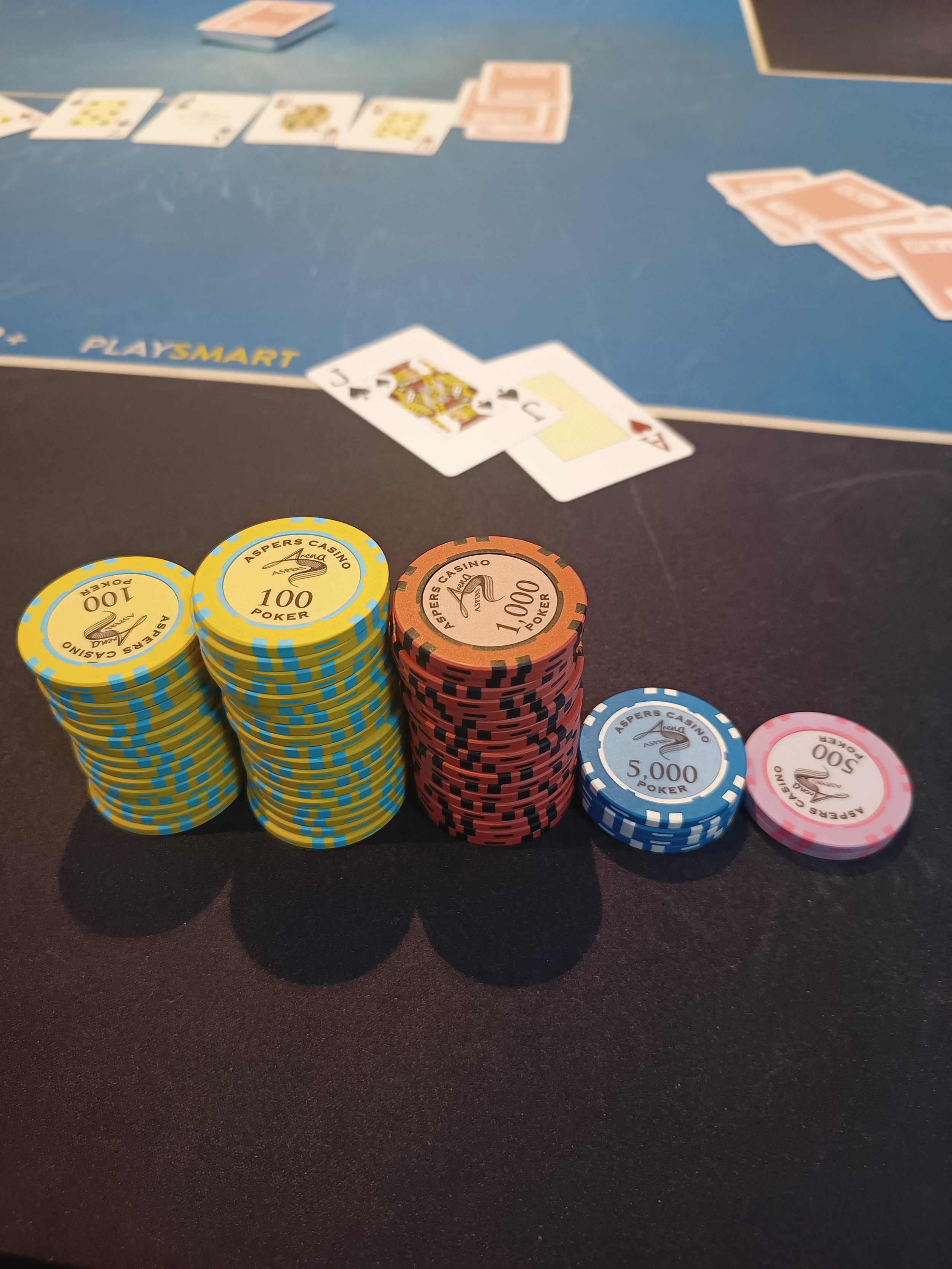 Stacks of poker chips with denominations from 100 to 5000, arranged on a black table. In the background, a blackjack game with playing cards and chips is visible.