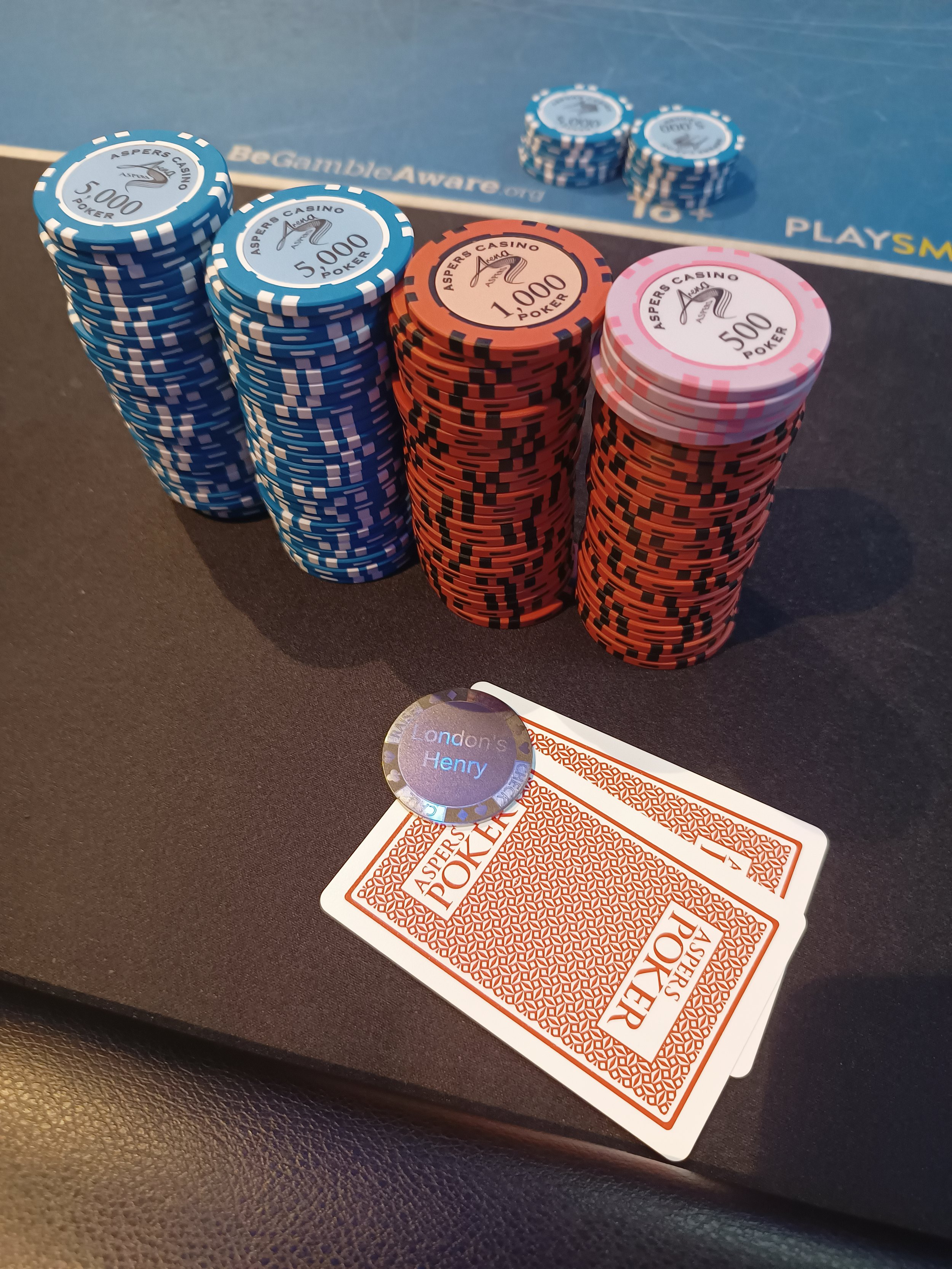 Stacks of poker chips in blue, orange, and pink colors with denominations of 5,000, 1,000, and 500, placed on a blackjack table with a pair of face-up playing cards and a poker chip nearby.