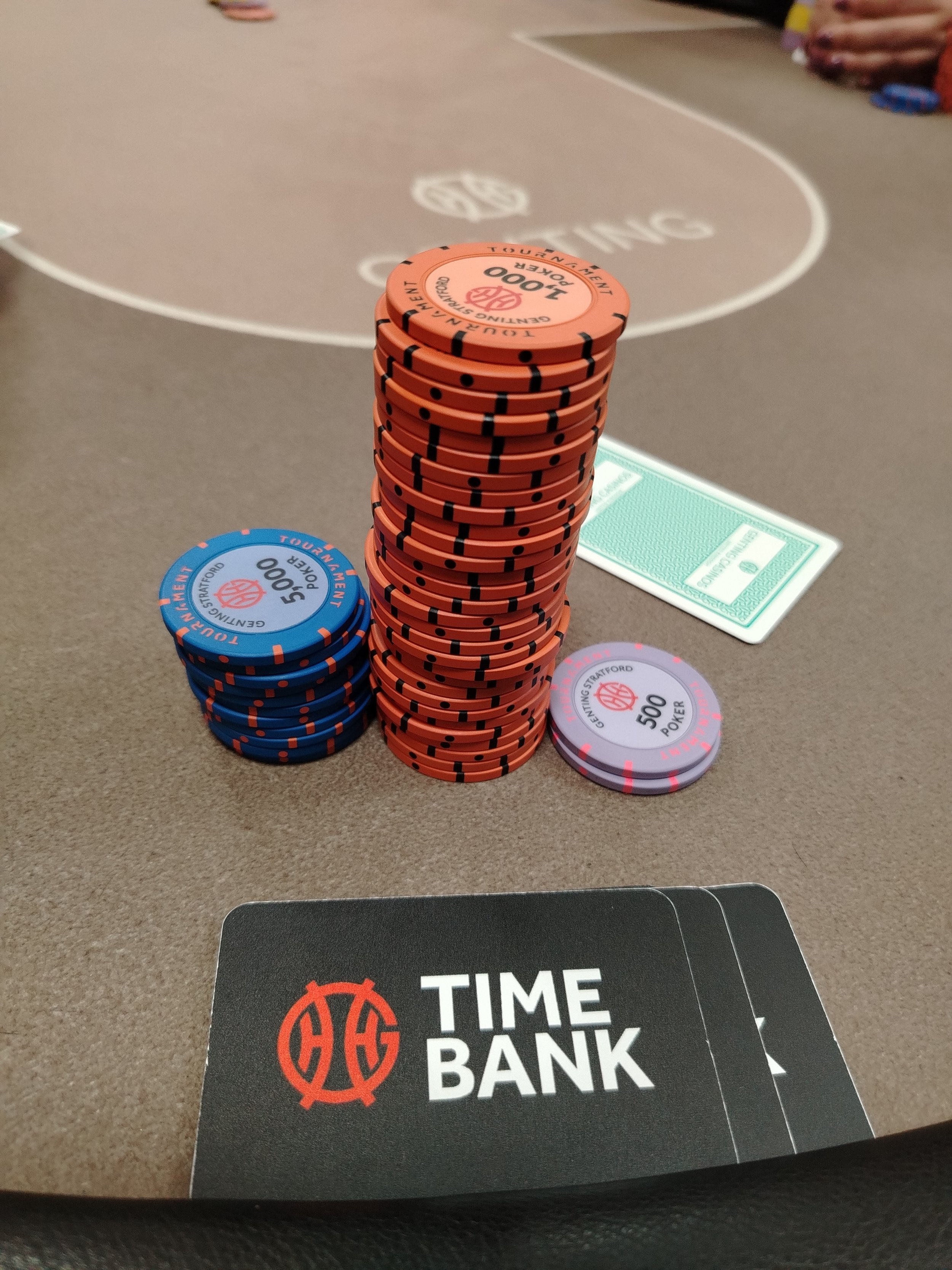 A stack of poker chips on a blackjack table in a casino or card room, with chips in orange, blue, and purple. A black card mat with red and white text reading 'TIME BANK' is in the foreground.