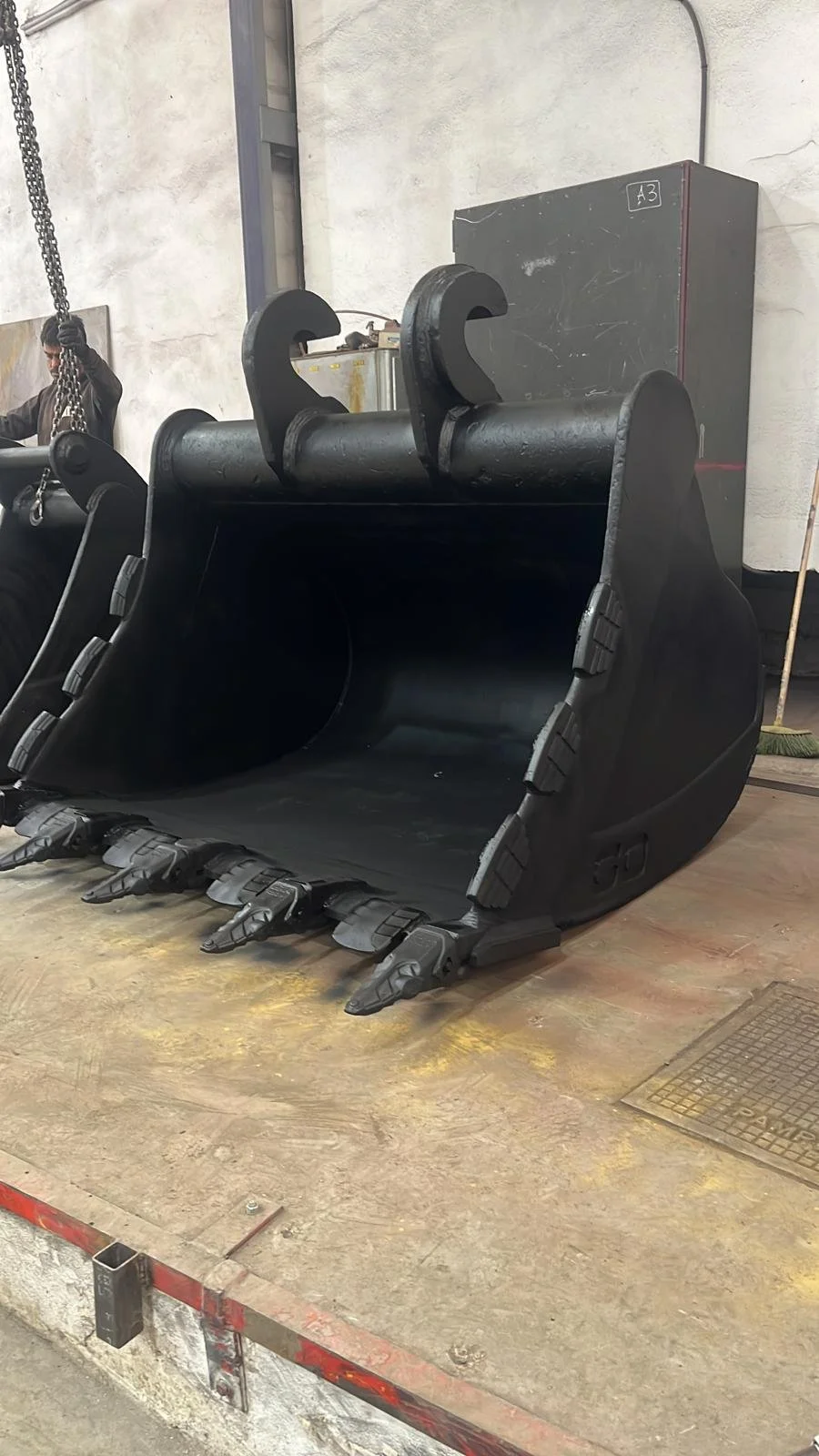 Imagen de un balde de excavadora grande en un taller o almacén, sin conductor, con detalles de un lugar industrial y una persona al fondo manejando una cadena.