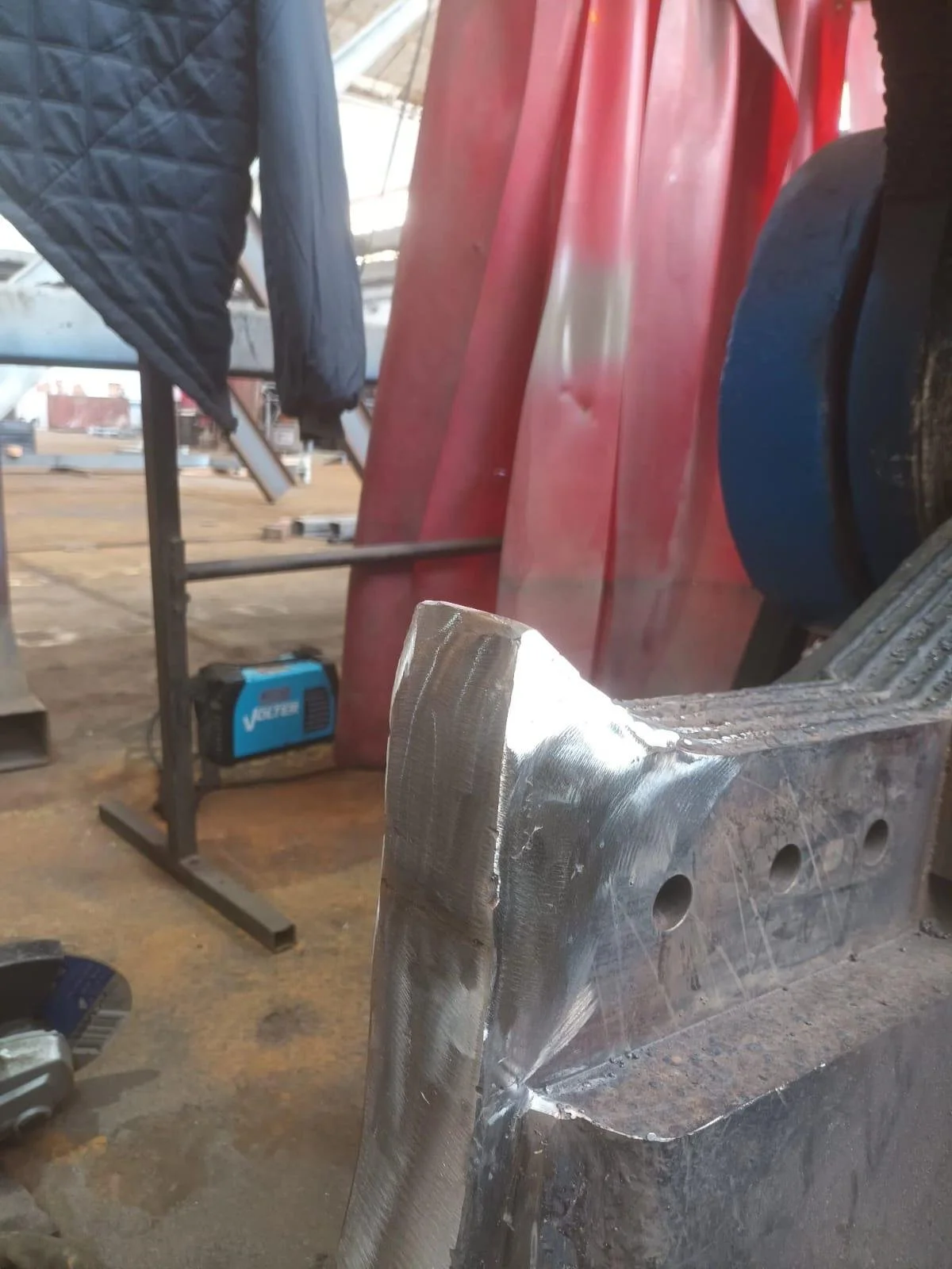 Imagen de un área de taller de soldadura con una pieza de metal en proceso de soldadura, con herramientas y equipos de protección en el fondo.